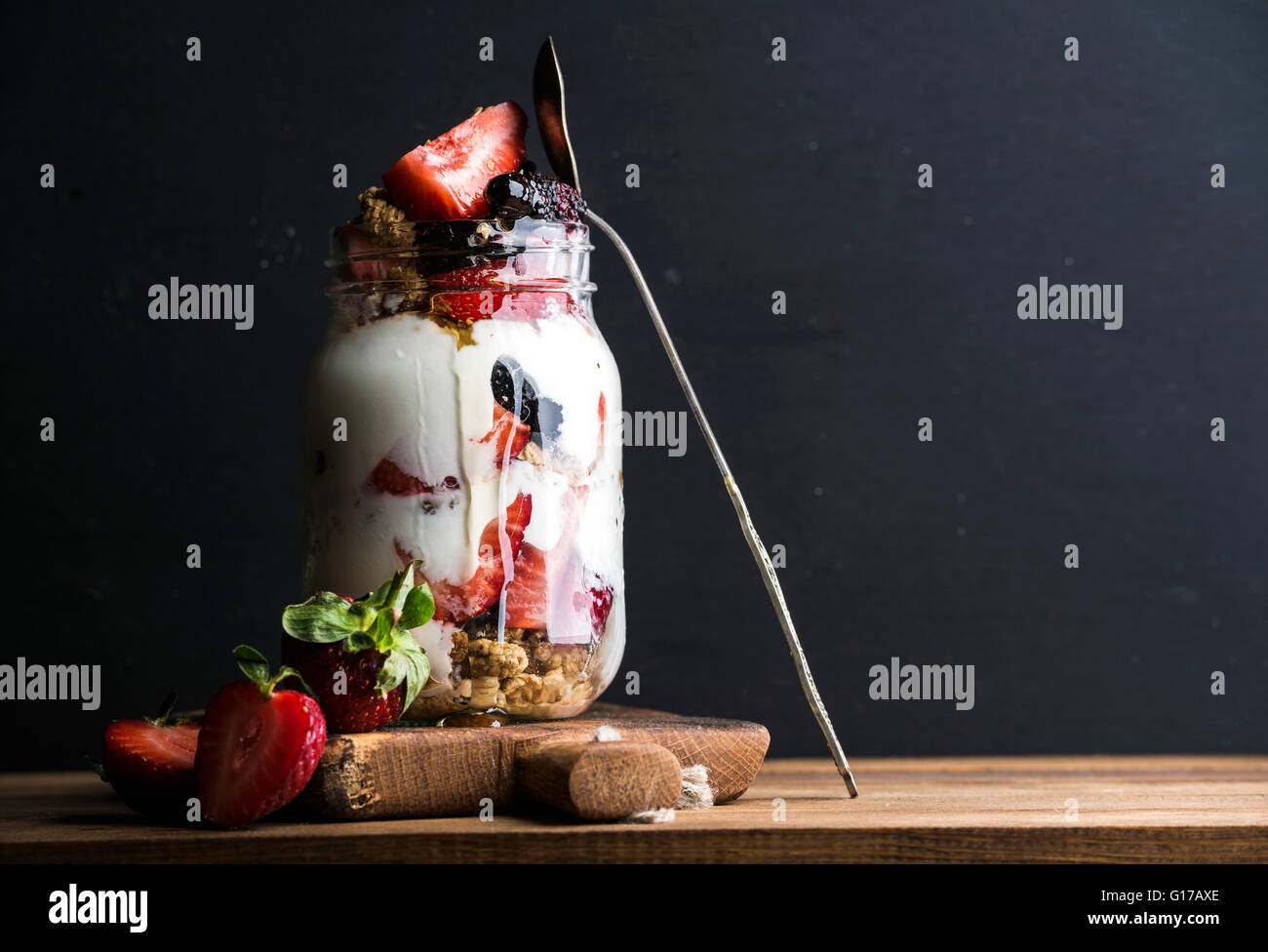 Joghurt-Hafer-Müsli mit Erdbeeren, Maulbeeren, Honig und Minze Blätter in Longdrinkglas Glas auf schwarzem Hintergrund, Tiefenschärfe, co Stockfoto