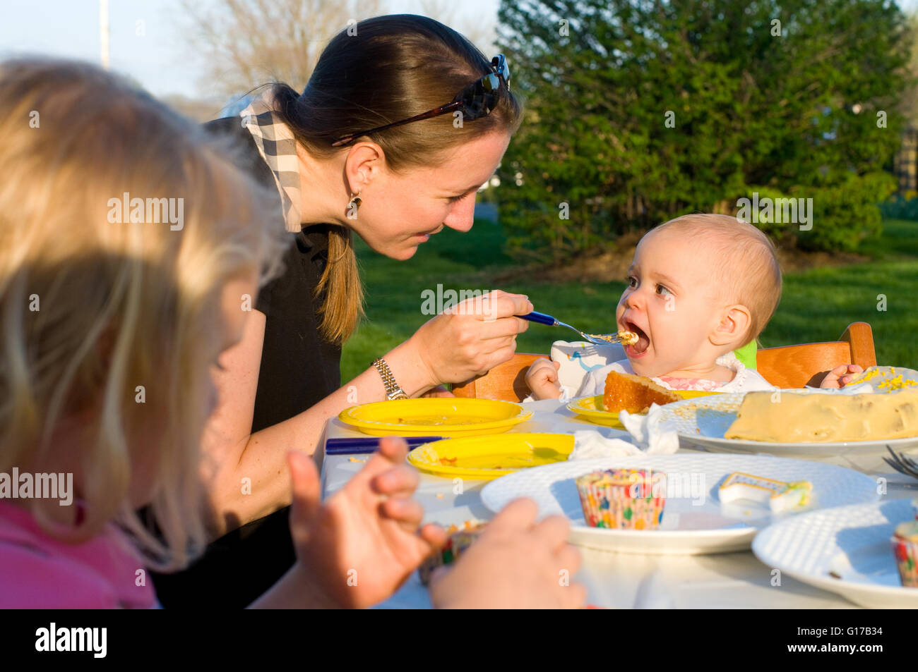Mutter und Kinder sitzen im Freien, Mutter Fütterung Baby Mädchen Geburtstagskuchen Stockfoto