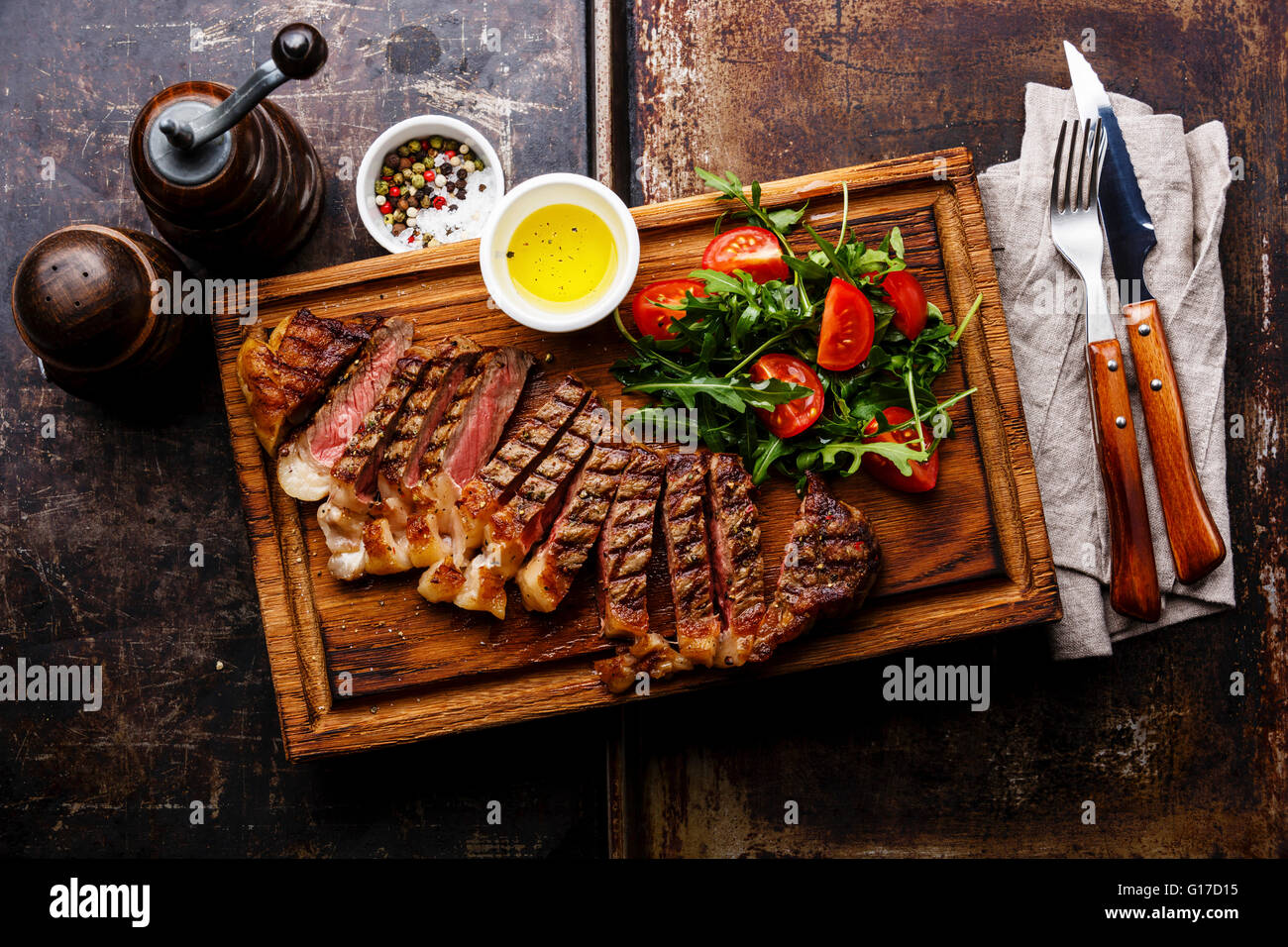 In Scheiben geschnittene gegrilltes Rindfleisch Grill Striploin Steak und Salat mit Tomaten und Rucola auf Schneidebrett auf dunklem Hintergrund Stockfoto