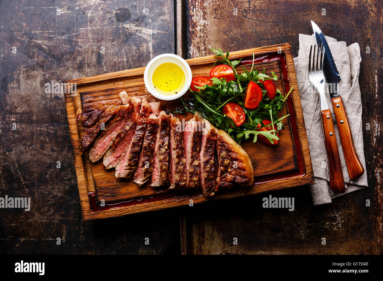 In Scheiben geschnittene gegrilltes Rindfleisch Grill Striploin Steak und Salat mit Tomaten und Rucola auf Schneidebrett auf dunklem Hintergrund Stockfoto