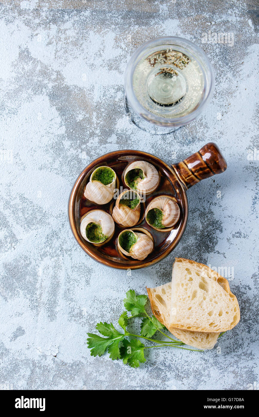 Escargots de Bourgogne - Schnecken mit Kräuter-Butter, in traditionellen keramischen Pfanne mit Petersilie, Brot und ein Glas Weißwein über b Stockfoto