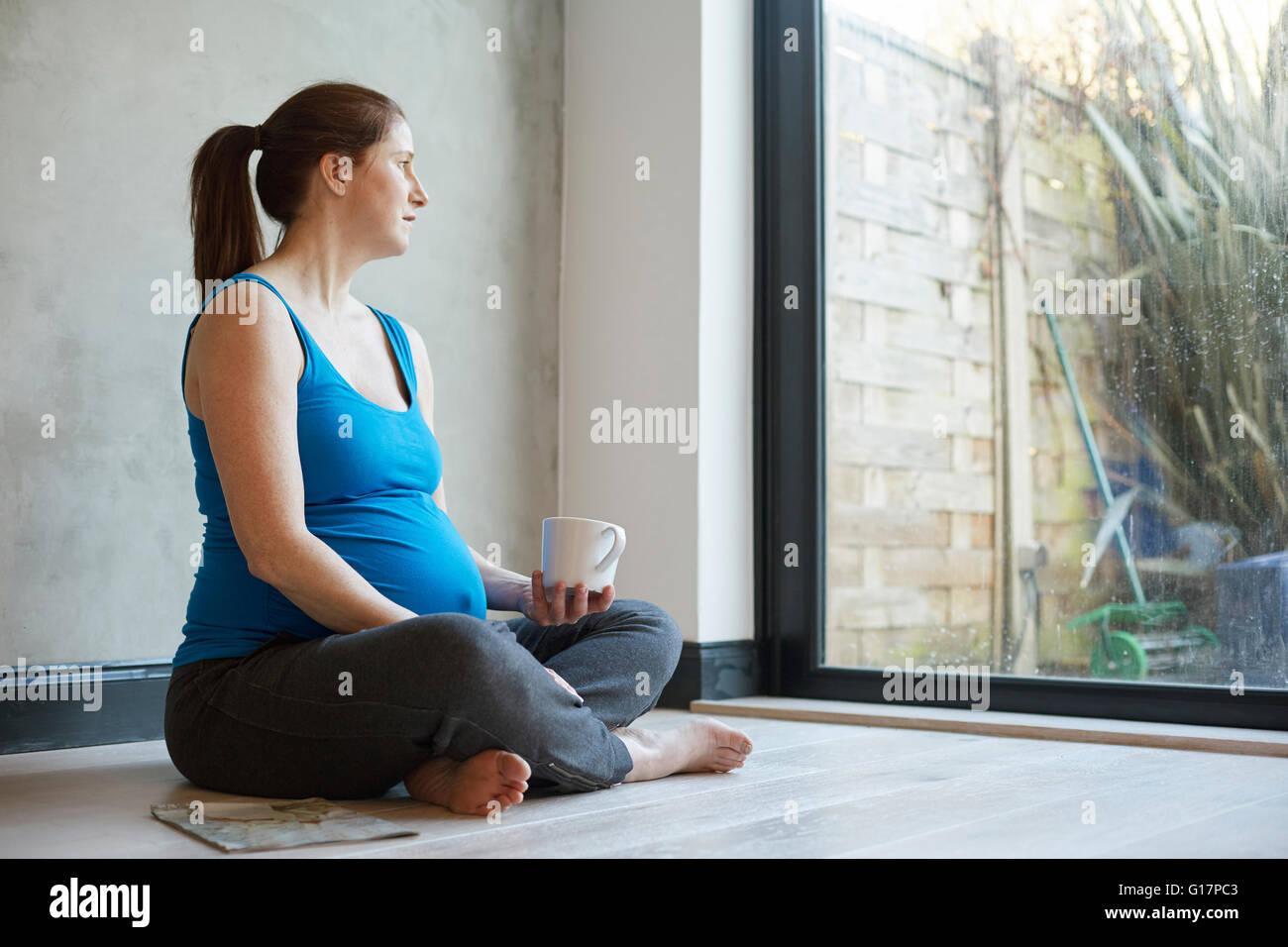 Schwangere Frau sitzen auf Boden überquerte Beinen halten Kaffee Tasse wegschauen aus Fenster Stockfoto