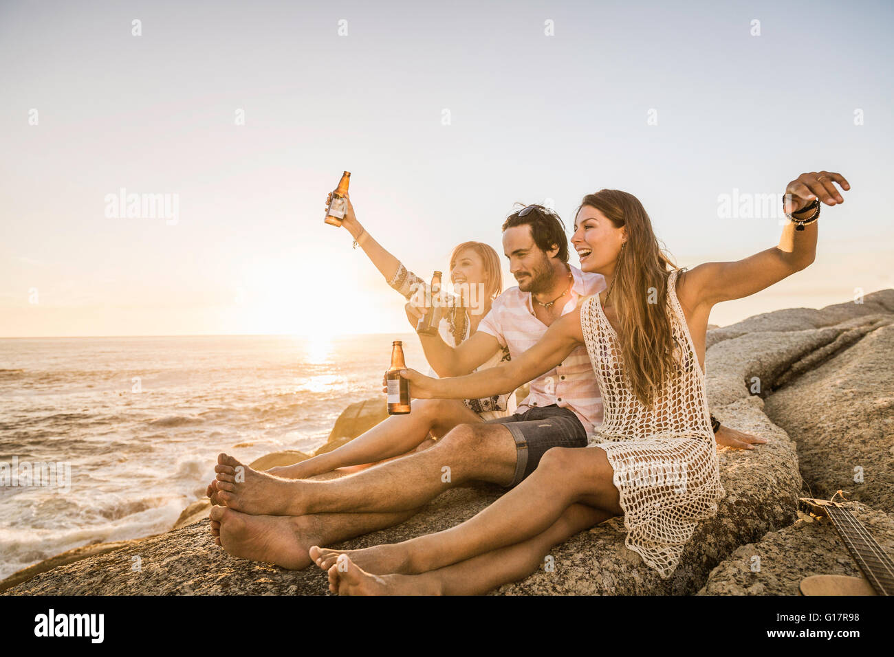 Drei Mitte Erwachsenen Freunde sitzen am Strand bei Sonnenuntergang Anhebung einen Toast, Cape Town, Südafrika Stockfoto
