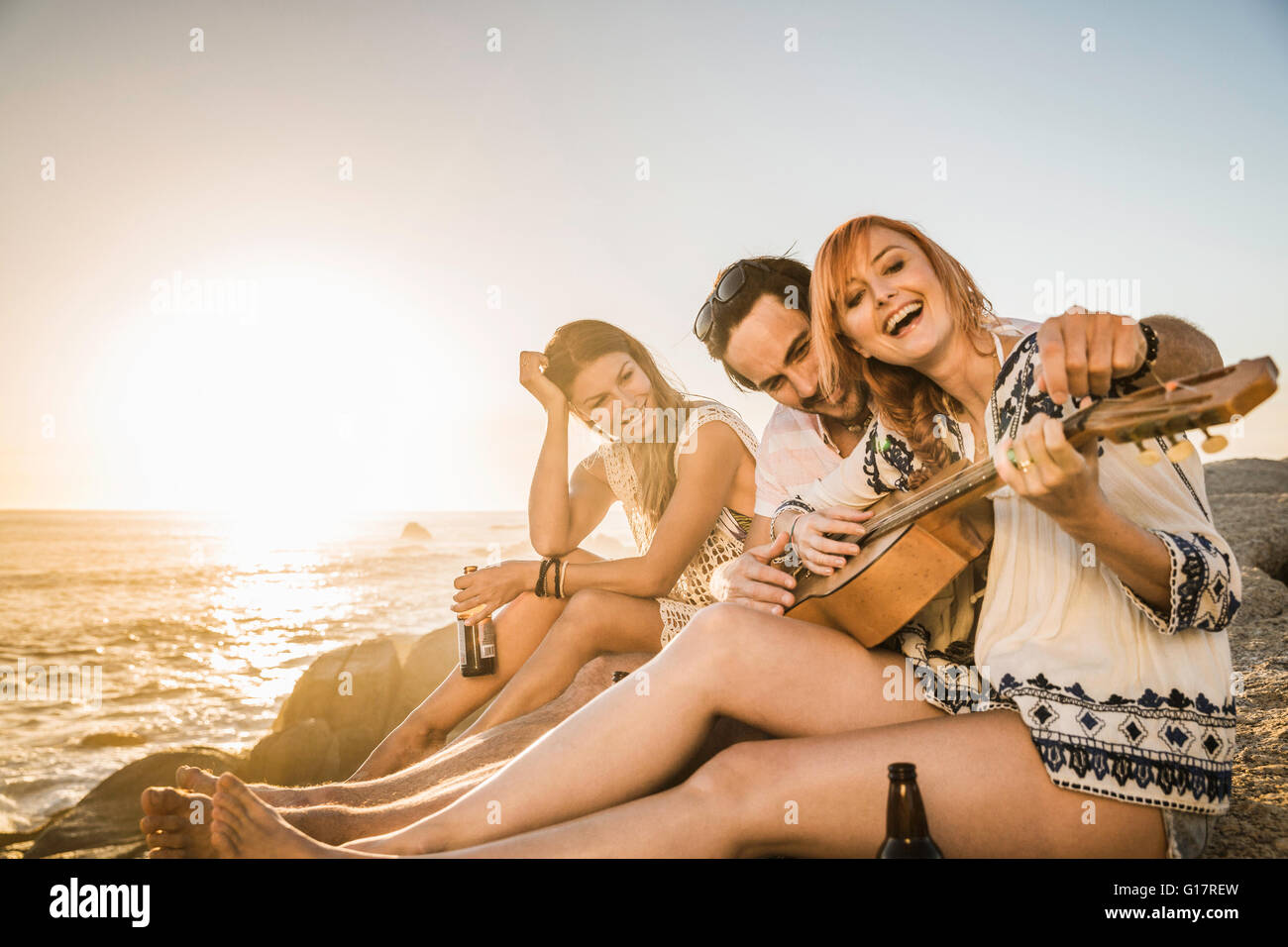 Drei Mitte Erwachsene sitzen am Strand spielt Gitarre bei Sonnenuntergang, Cape Town, Südafrika Stockfoto