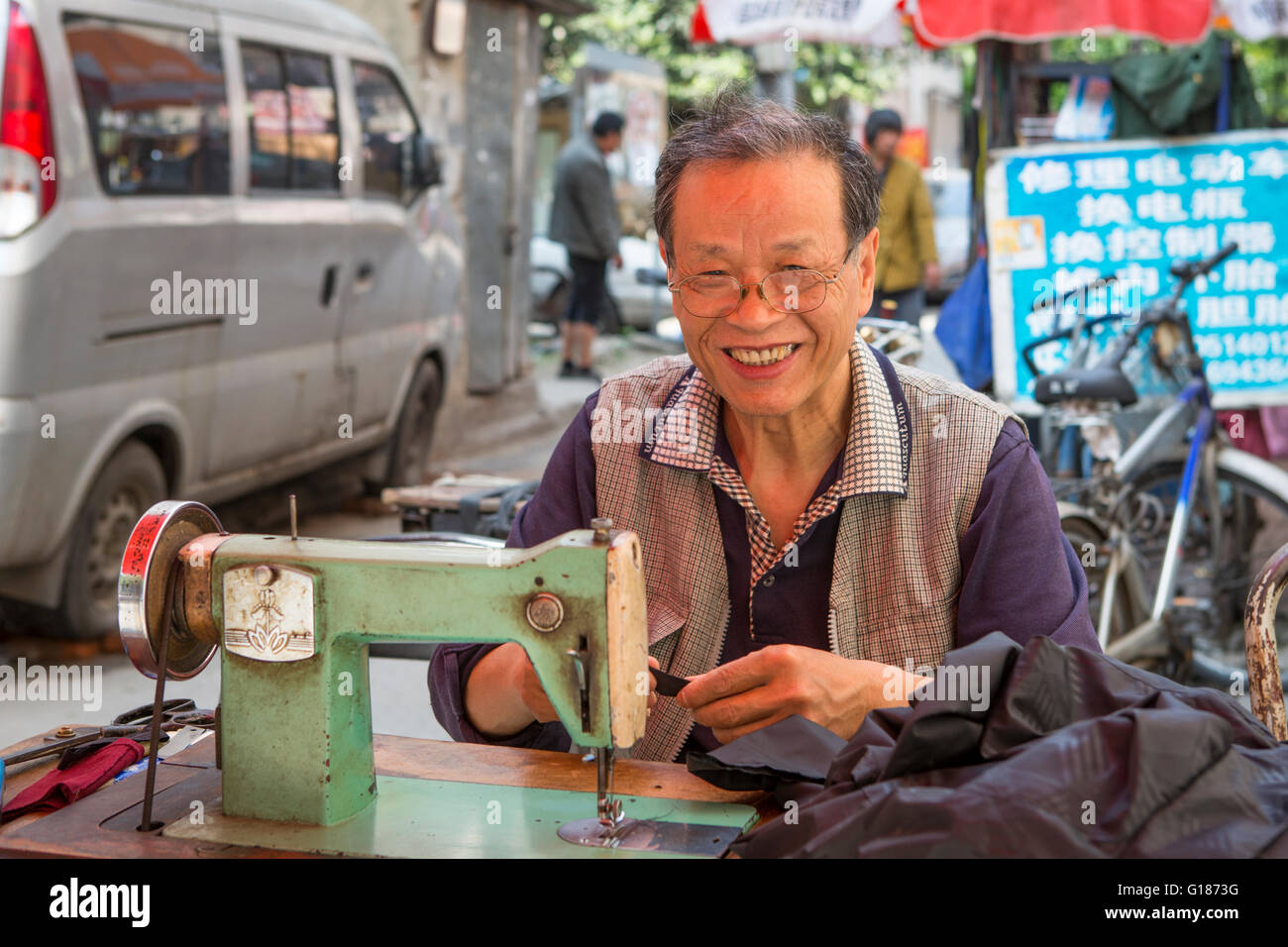 Lächelnde chinesische senior Schneider Unternehmer Nähen im Freien auf einer alten Nähmaschine in einer Straße in Nanjing / China Stockfoto