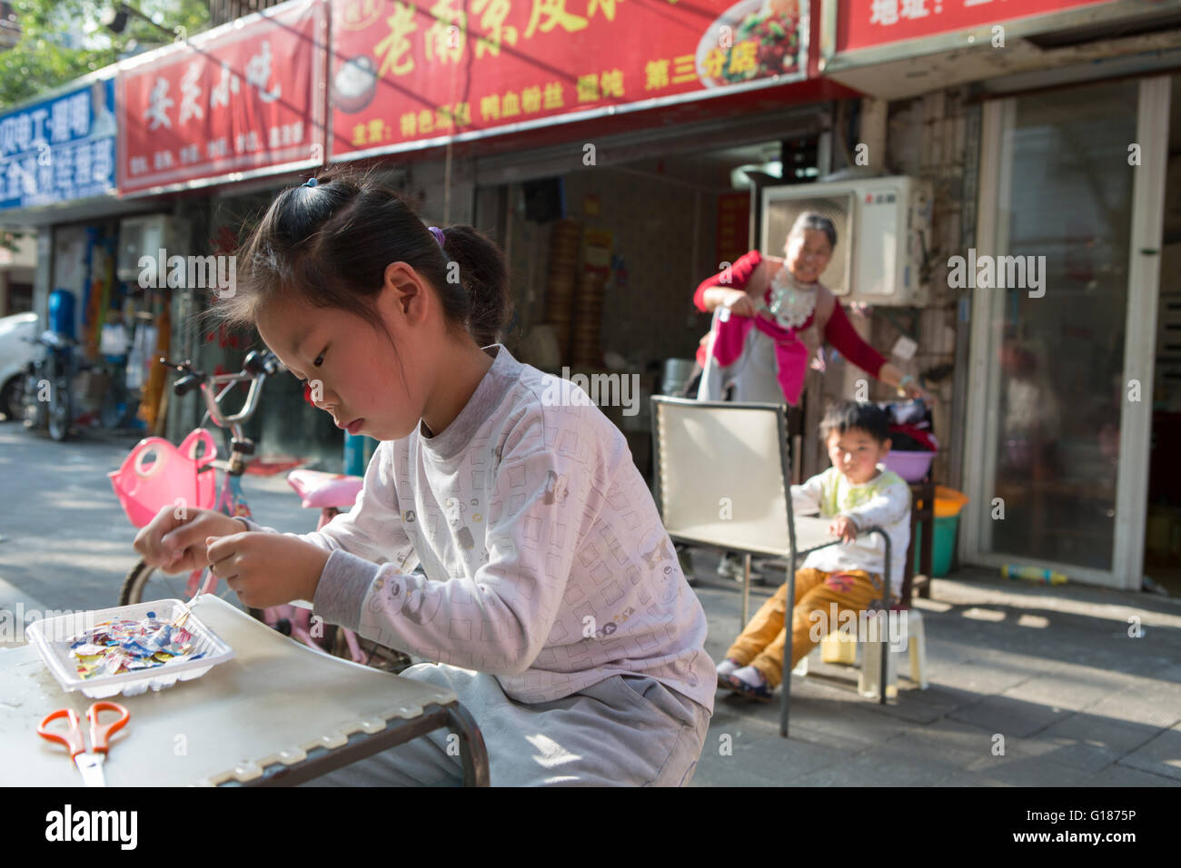 Chinesisches Mädchen spielen vor ihrem Haus auf dem Bürgersteig einer Straße in Nanjing mit ihrem Bruder und ihrer Mutter, die gerade im freien Stockfoto