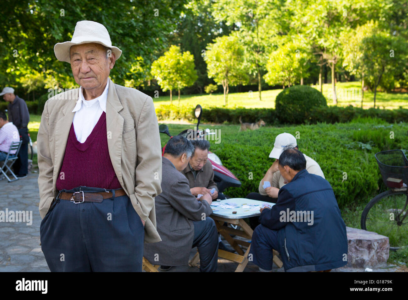 Porträt eines chinesischen Mann mit einem weißen Hut in einem Park mit Menschen Spielkarten im Hintergrund an einem sonnigen Tag Stockfoto