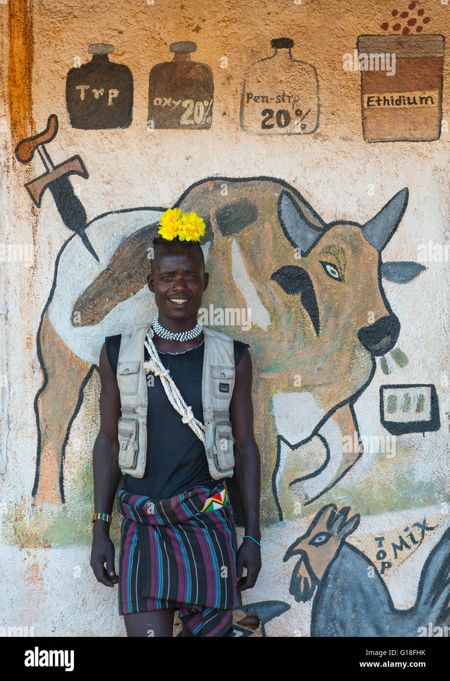 Lächelnd Bana Stamm Mann mit gelben Kunststoff Blume im Haar vor ein Wandbild Darstellung einer Kuh, Omo-Tal, Key Afer, Ethiopi Stockfoto
