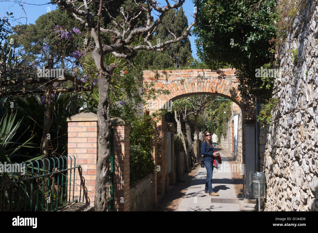 Eine weibliche Touristin unter einem Backsteinbogen und Einheimische in der ruhigen Fußgängerzone Via Tragara auf der Insel Capri im Golf von Neapel, Italien Stockfoto