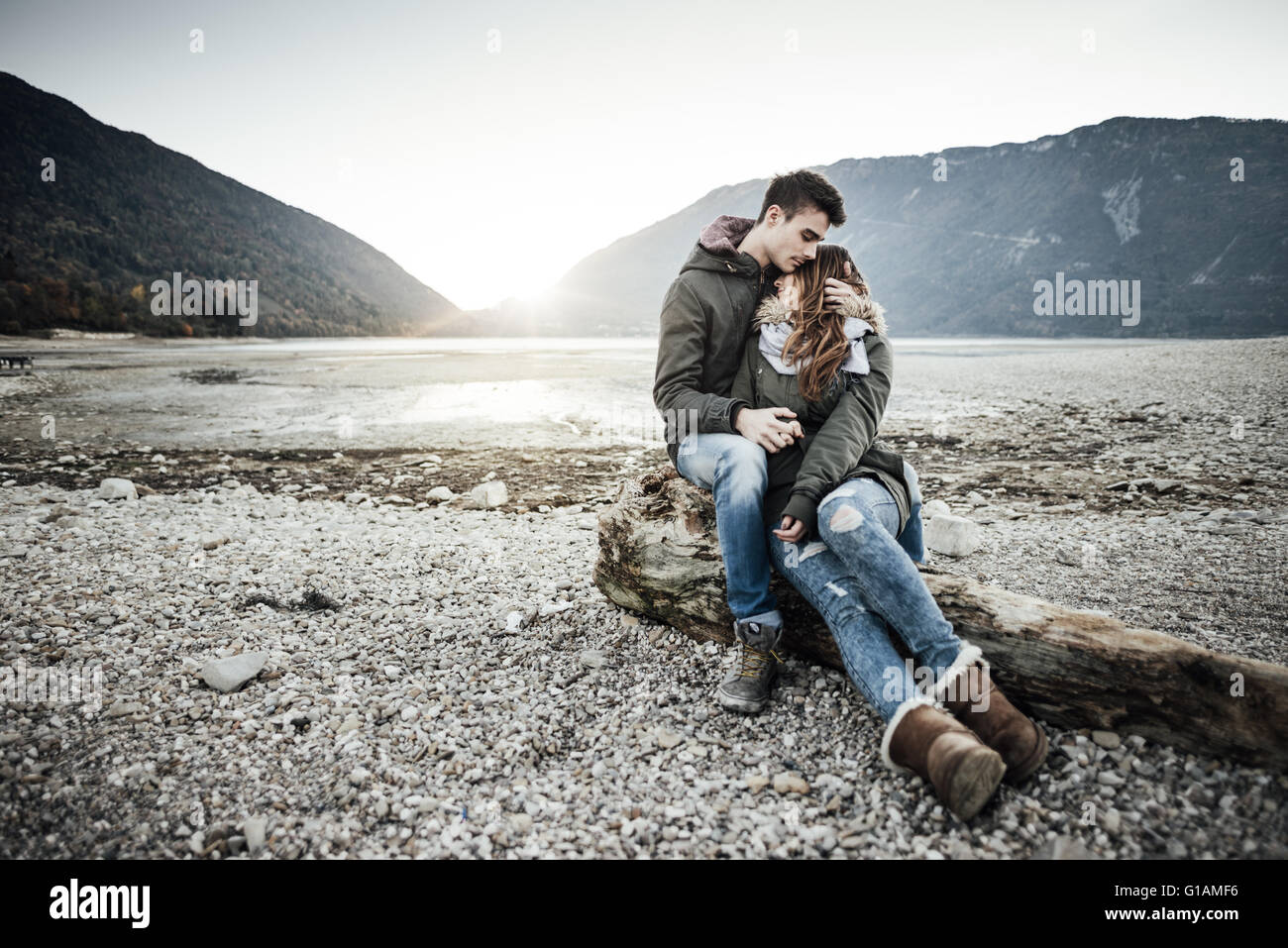 Junges Paar mit einem romantischen Date am See, sie umarmen und sitzen auf einem Baumstamm am Strand, Liebe und Beziehungen Stockfoto