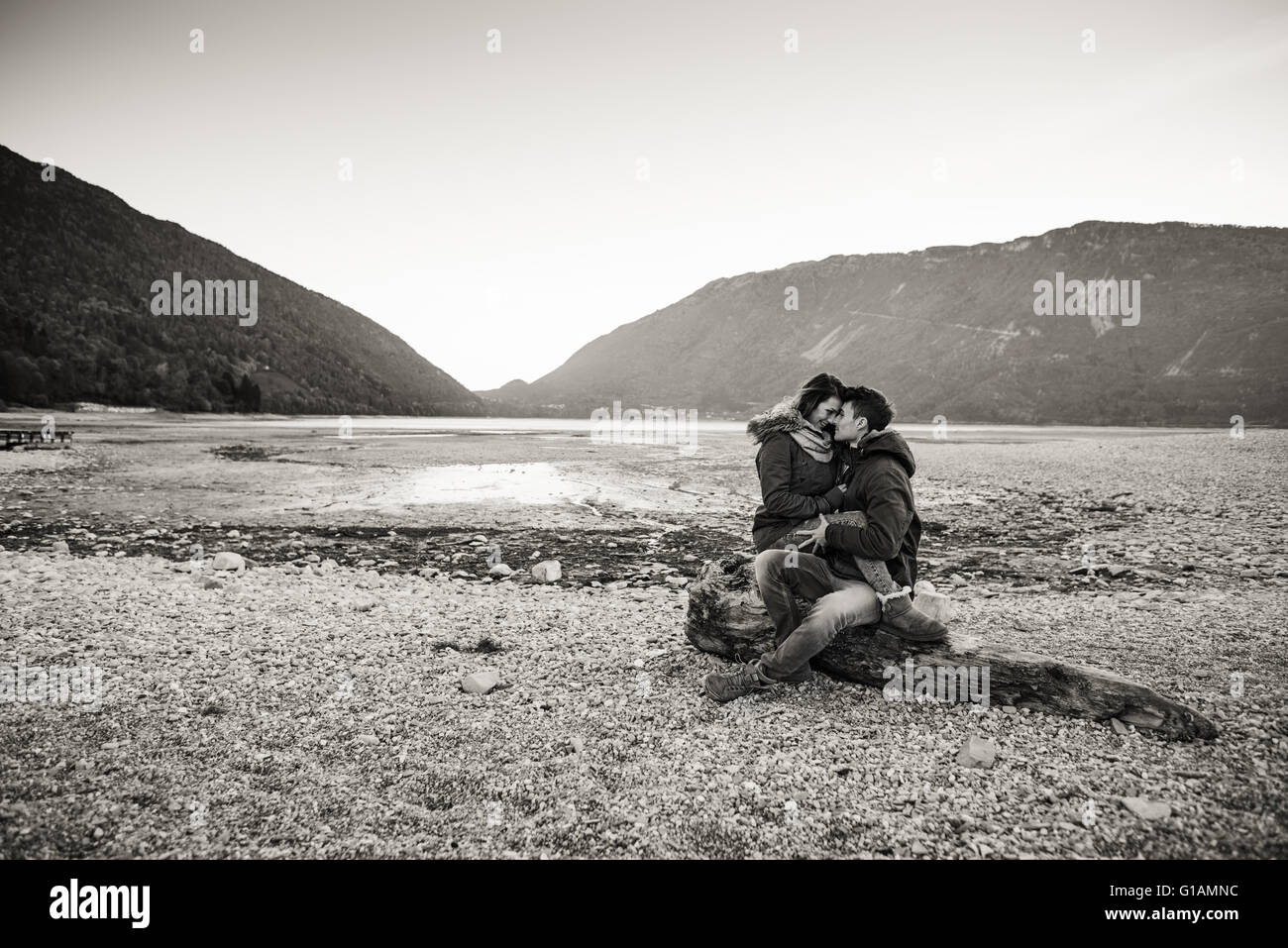 Junges Paar mit einem romantischen Date am See, sie umarmen und sitzen auf einem Baumstamm auf dem Strand, Liebe und Beziehungen co Stockfoto