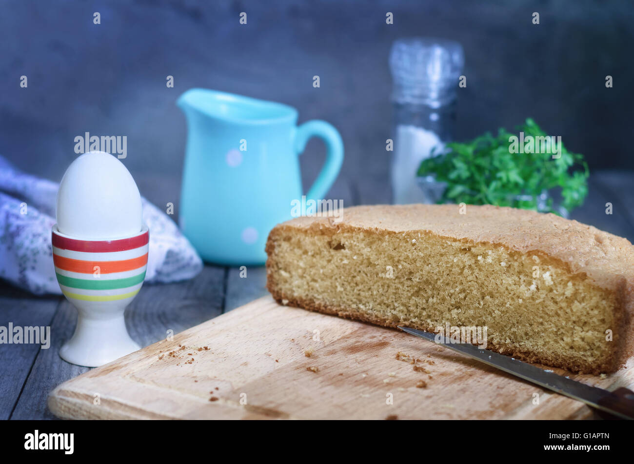 Die Hälfte des Brotes auf ein Brett, Messer und Ei in einem gestreiften Stand. Im Hintergrund: blauer Krug, Salzstreuer, Serviette und Bunc Stockfoto