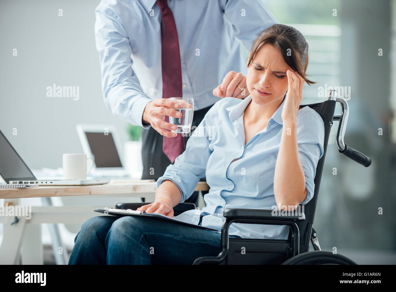 Business-Frau im Rollstuhl mit einer Kopfschmerzen im Büro, ihr Kollege ist ihr ein Glas Wasser und ihr zu helfen Stockfoto