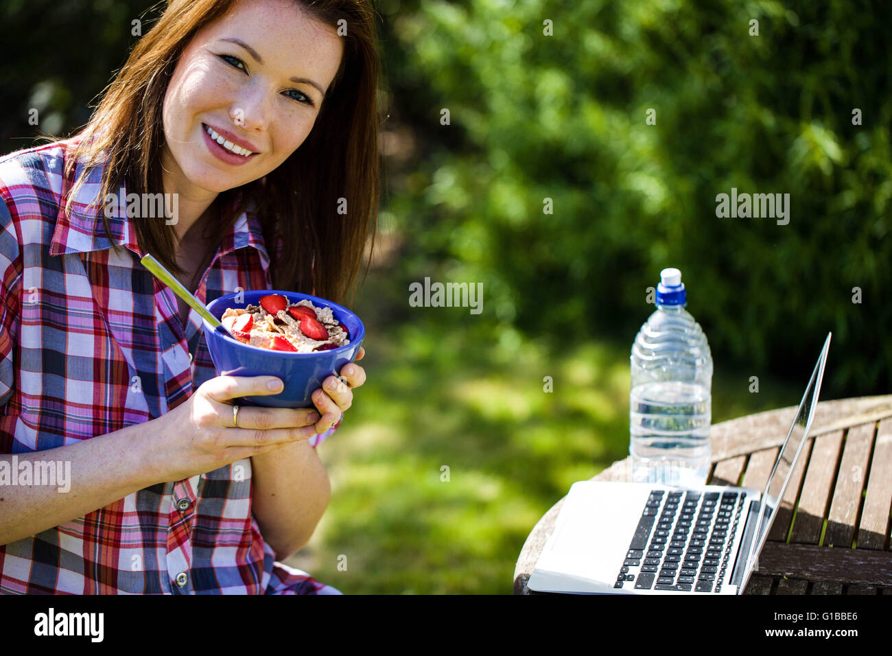 Porträt einer glücklichen Frau Essen Frühstückskost aus Getreide außerhalb In einem Garten mit ihrem Laptop und eine Flasche Wasser Stockfoto