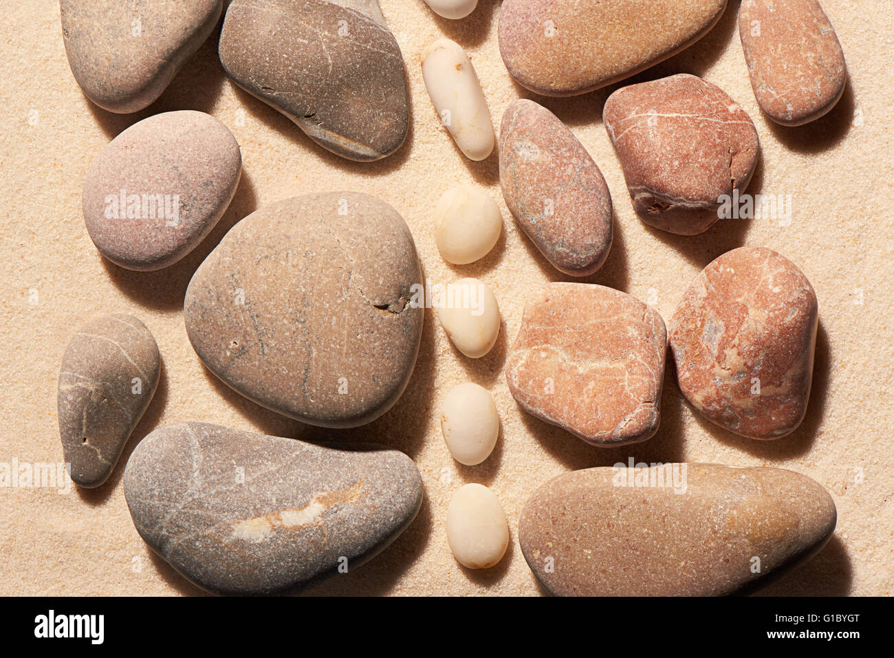 Sammlungen von rosa und grau getönten Meer Steinen durch vertikalen Streifen von kleineren weißen Steinen auf Sand getrennt. Sommerstrand Stockfoto