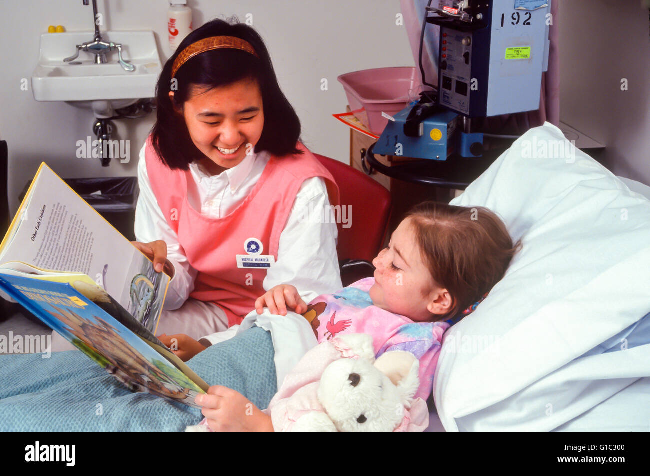 Kind helfen, ein anderes Buch lesen Teenager im Alter von 16 und 17 Jahren koreanisch-amerikanischen Candystripper freiwillige Bett von sehr kranken Mädchen 6-7 Jahre alten © Myrleen Pearson Stockfoto