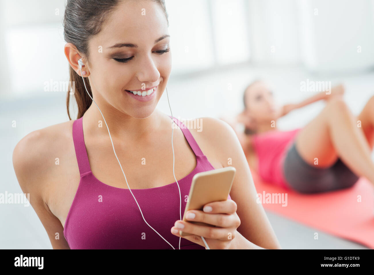 Junge lächelnde Frau im Fitnessraum entspannen und Musikhören mit einem Mobiltelefon und Kopfhörer Stockfoto