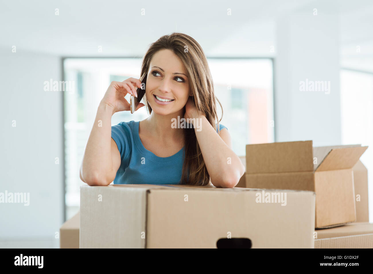 Schöne junge Frau mit einem Anruf in ihrem neuen Haus, sie lehnt sich auf einen Karton Stockfoto