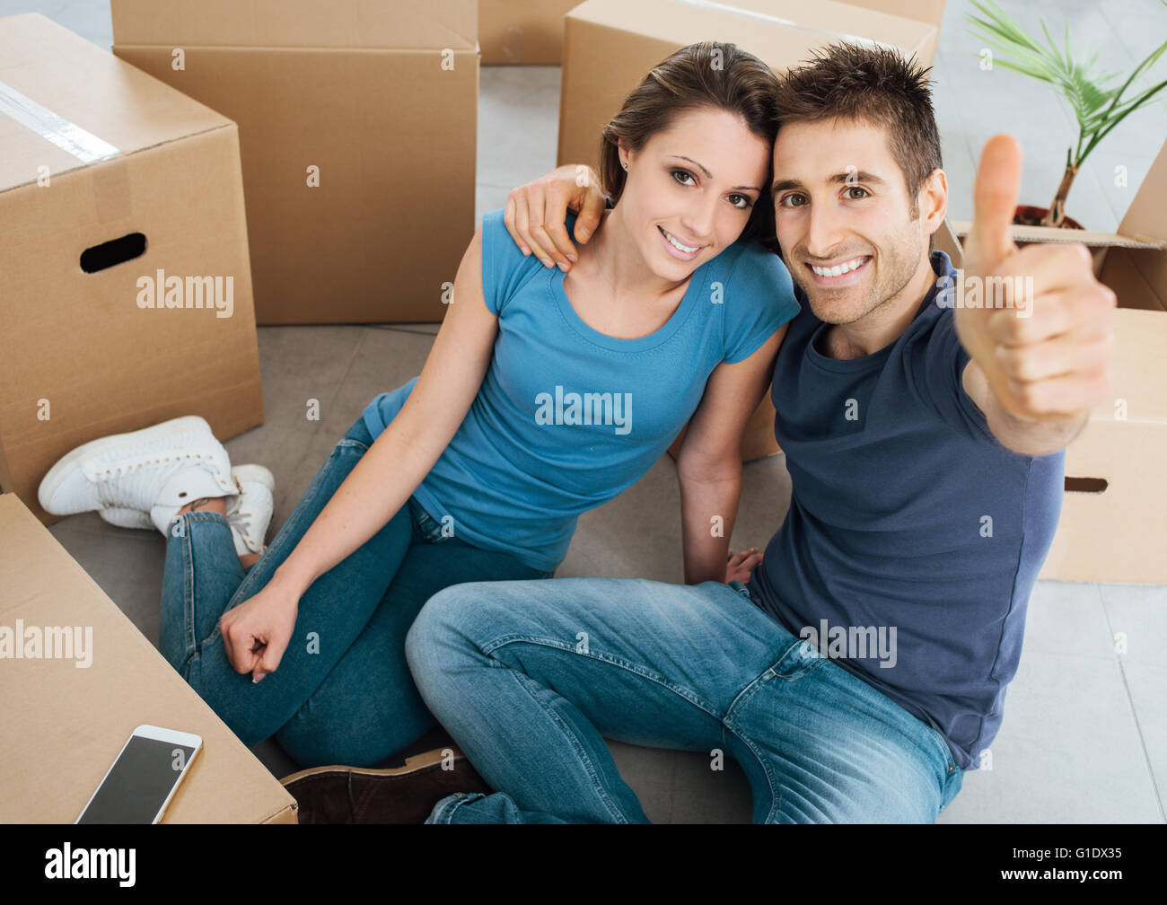 Glückliches junges Paar Daumen auf und lächelt in die Kamera, sitzen sie auf ihrem neuen Haus Boden umgeben von Kartons Stockfoto
