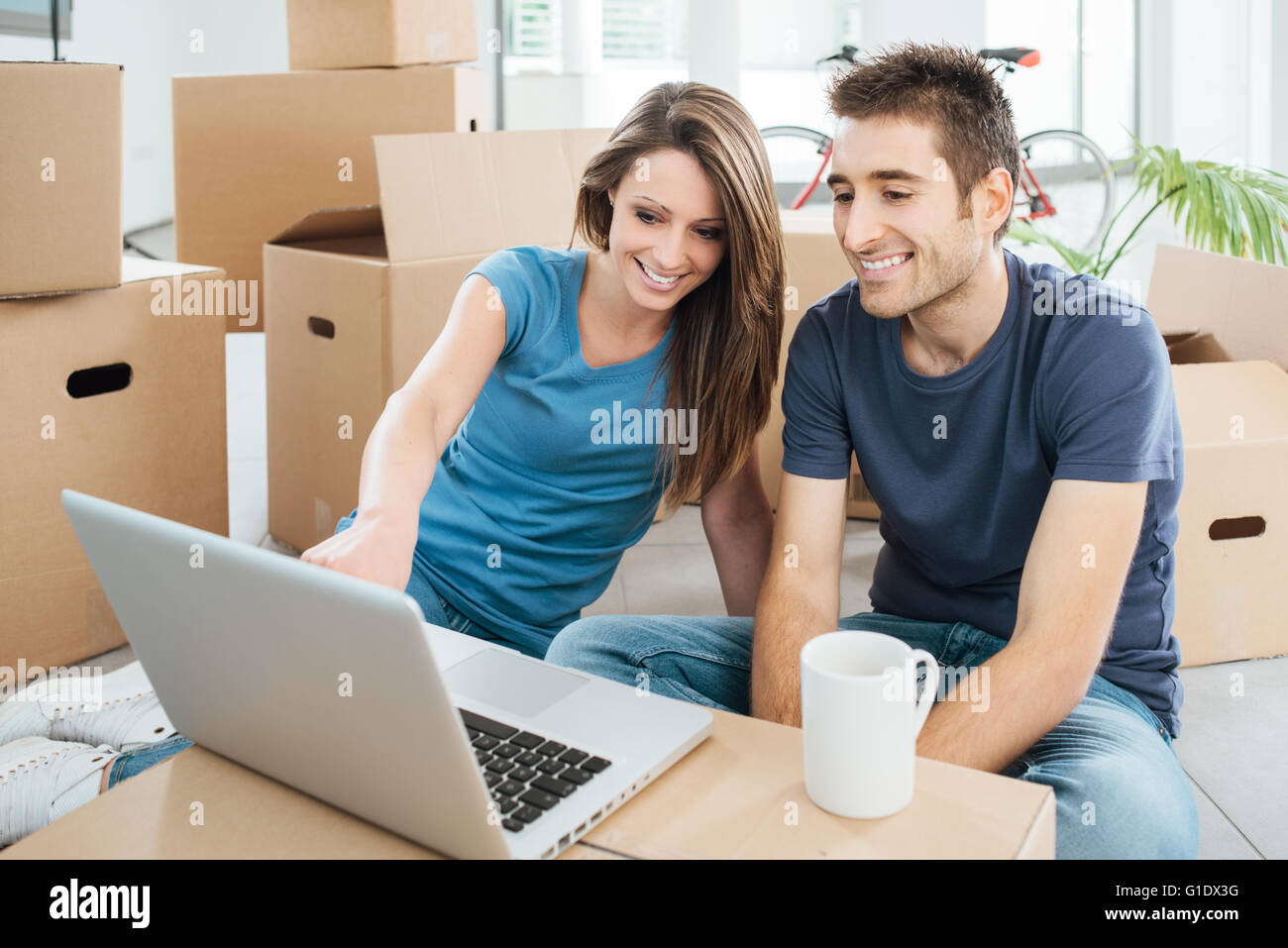 Brautpaar sitzt auf ihrem neuen Haus Boden umgeben von Kartons und eine drahtlose Laptop während einer Kaffeepause mit Stockfoto