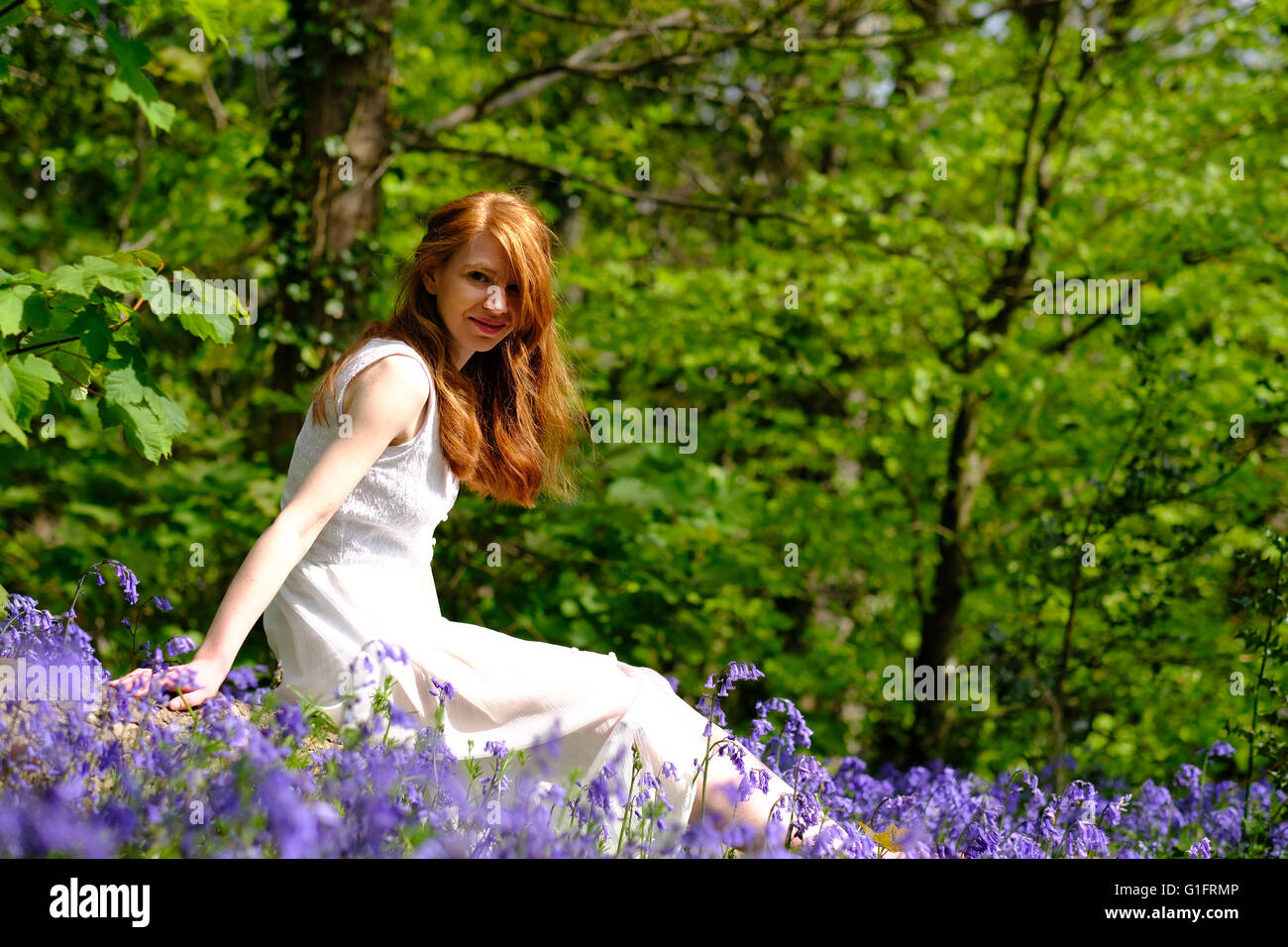 Eine schlanke, hellhäutige, junge Frau mit roten Haaren sitzt unter Bluebell Blumen im Wald Stockfoto