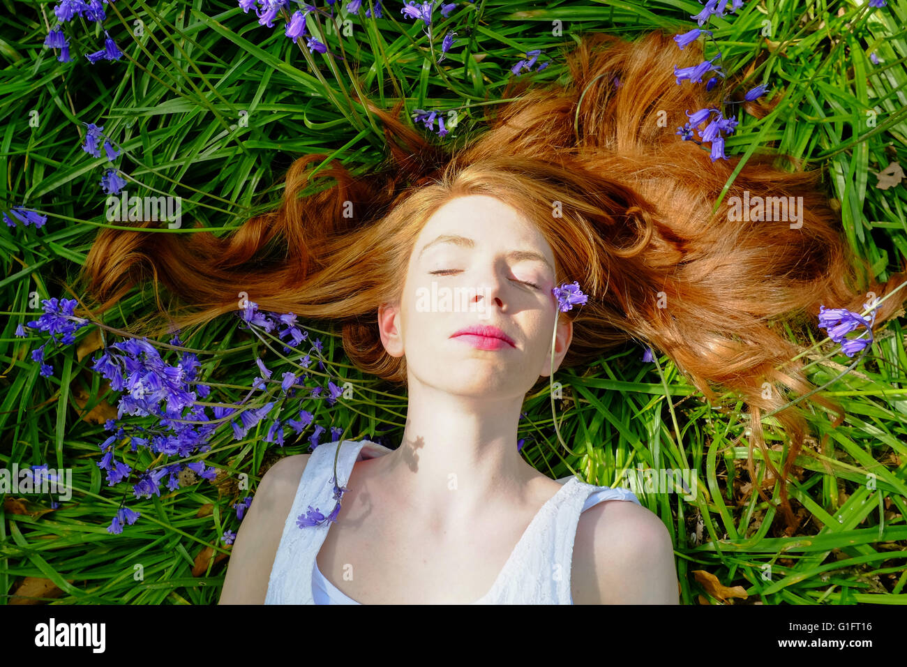 Eine hellhäutige junge Frau mit roten Haaren und roten Lippen liegt mit ihren Haaren zu verbreiten und unter Bluebell Blumen geschlossenen Augen. Stockfoto