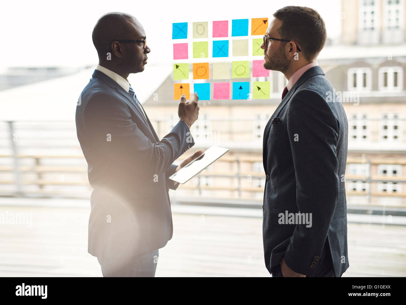 Zwei Geschäftsleute haben eine ernsthafte Diskussion, wie sie vor Reihen von bunten Memos auf eine Glasscheibe brainstorming eine n stehen Stockfoto
