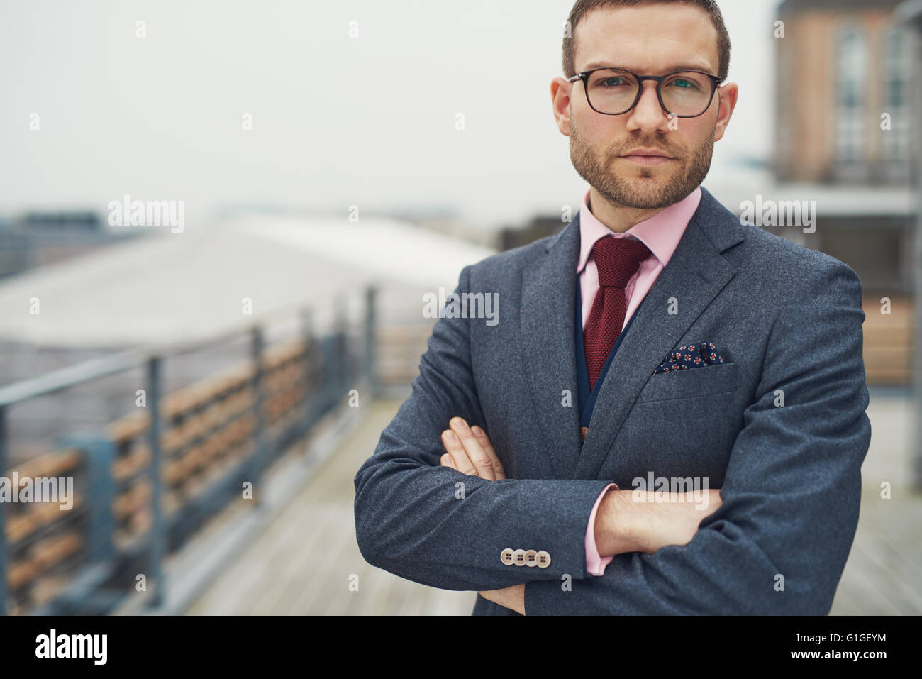 Stilvolle schwere hübscher bärtiger junger Geschäftsmann in Brillen, rote Krawatte und verschränkten Armen draußen am Balkon in der Nähe von Geländer Stockfoto