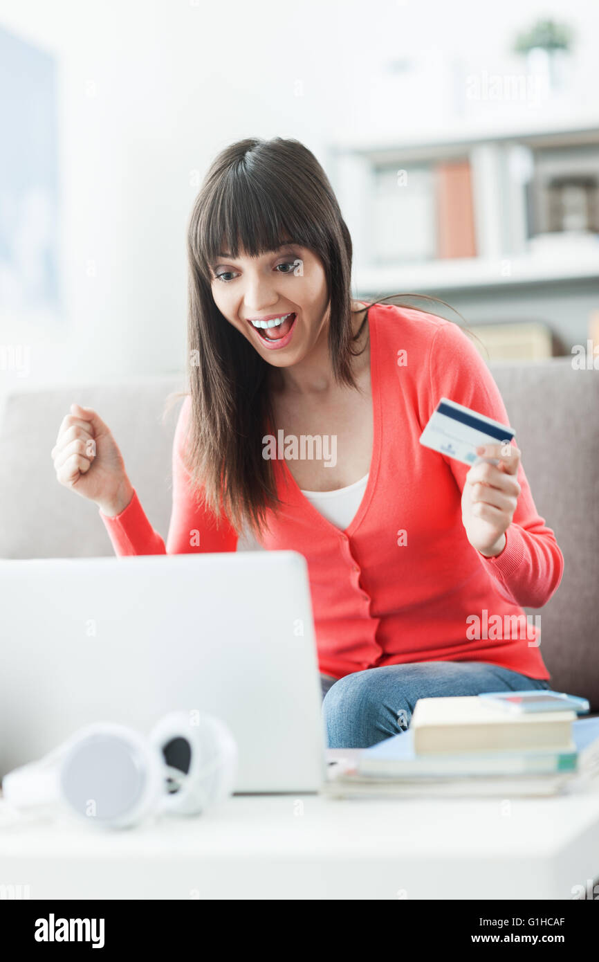 Lächelnde junge Frau zu Hause einen Laptop verwenden und Online-shopping mit Kreditkarte, ist sie fröhlich mit ein erhobener Faust Stockfoto