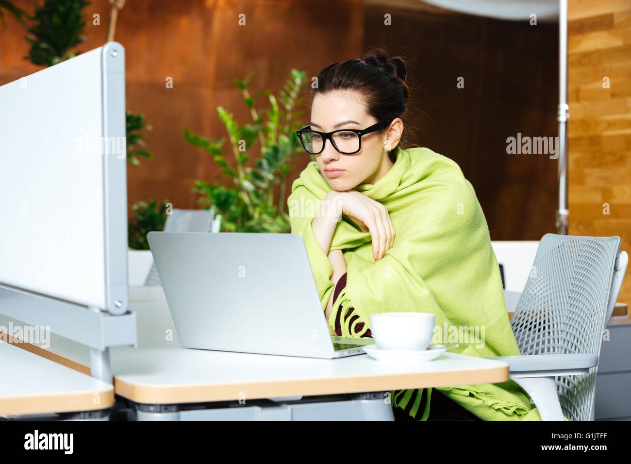 Schöne nachdenkliche junge Frau begehrt mit Tagesdecke mit Laptop und Kältegefühl am Arbeitsplatz Stockfoto