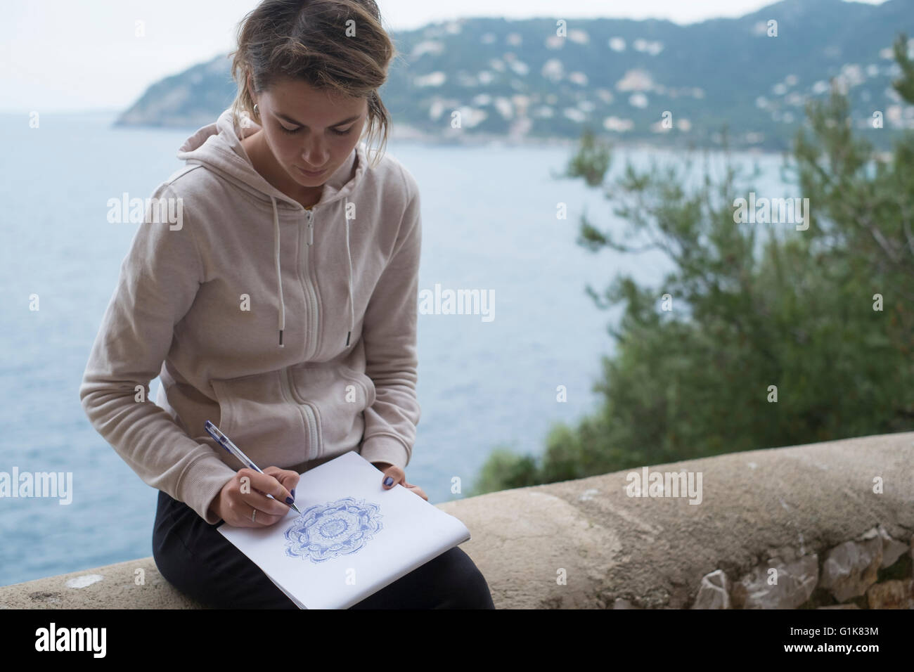 Junge Frau zeichnen eine Mandala-Muster auf einem Skizzenblock mit einem violetten Bleistift im Freien mit dem Meer hinter ihr Stockfoto