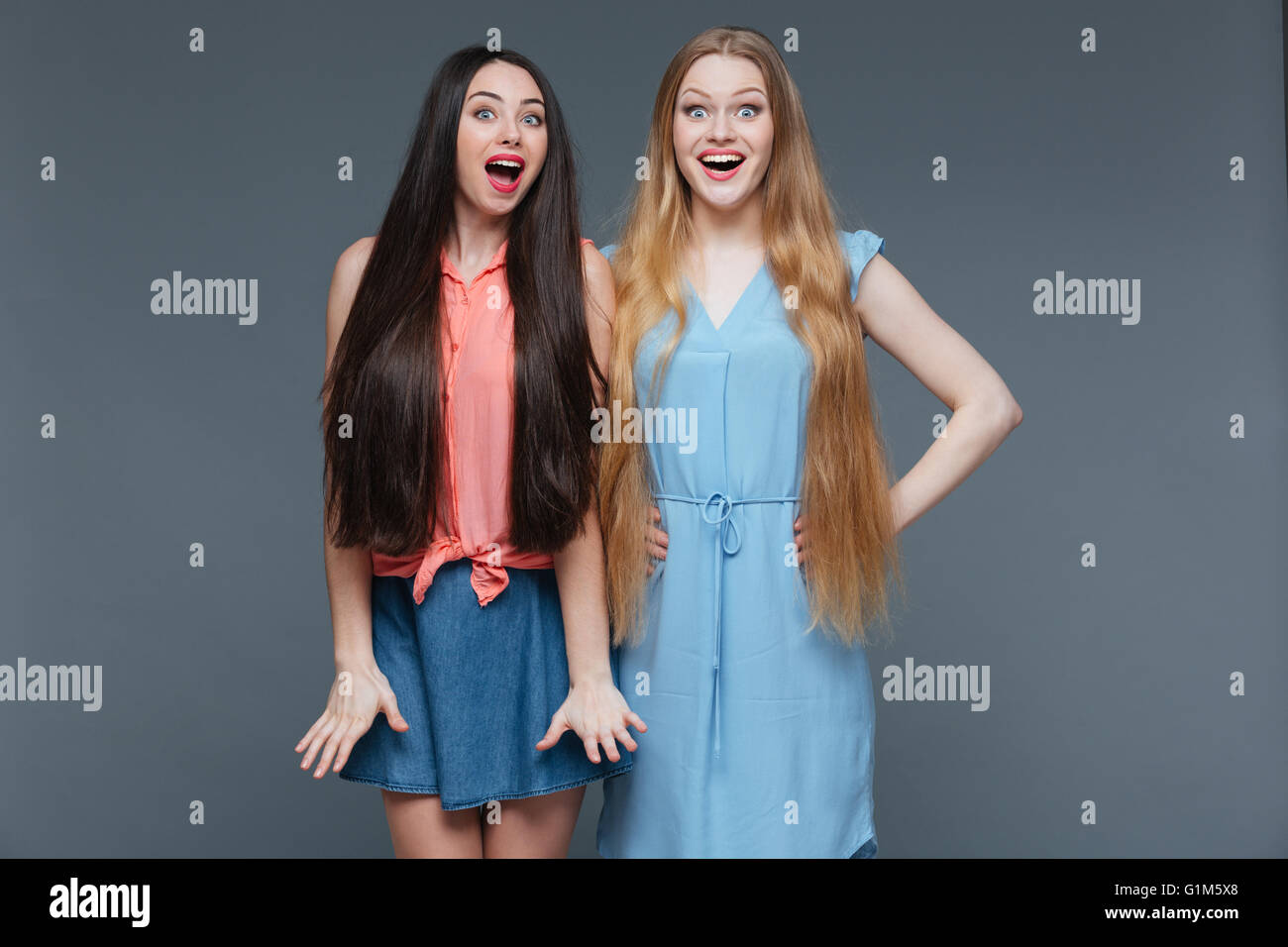 Zwei glückliche überraschte junge Frauen mit langen Haaren stehen mit Mund öffnete sich über grauen Hintergrund Stockfoto