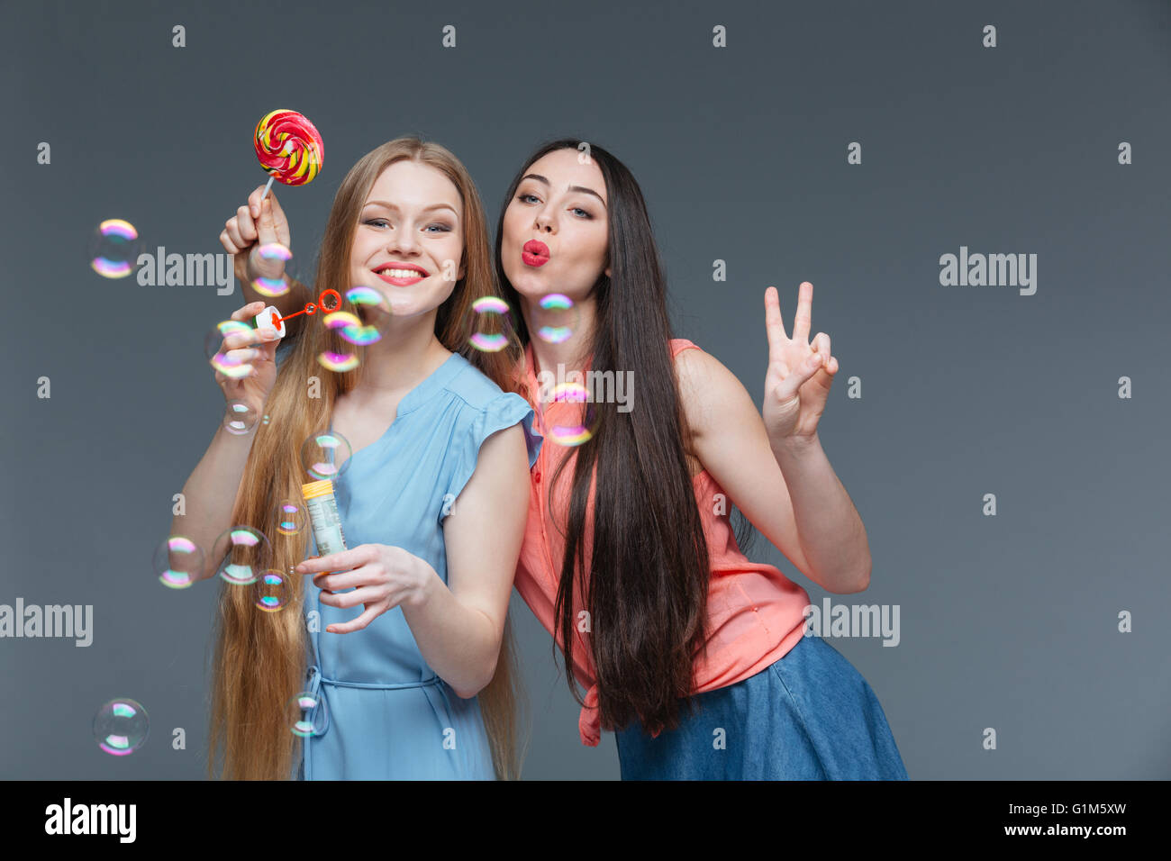 Zwei glückliche verspielt junge Frauen mit bunten Lollipop Seifenblasen auf grauem Hintergrund Stockfoto