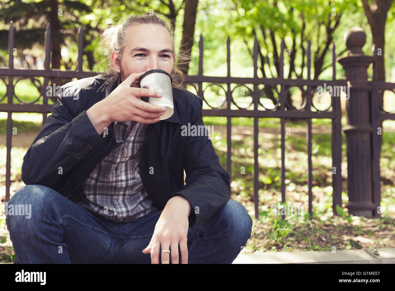 Bärtige asiatischen Mann sitzt auf dem Bürgersteig in Park und trinken Kaffee aus Pappbecher Stockfoto