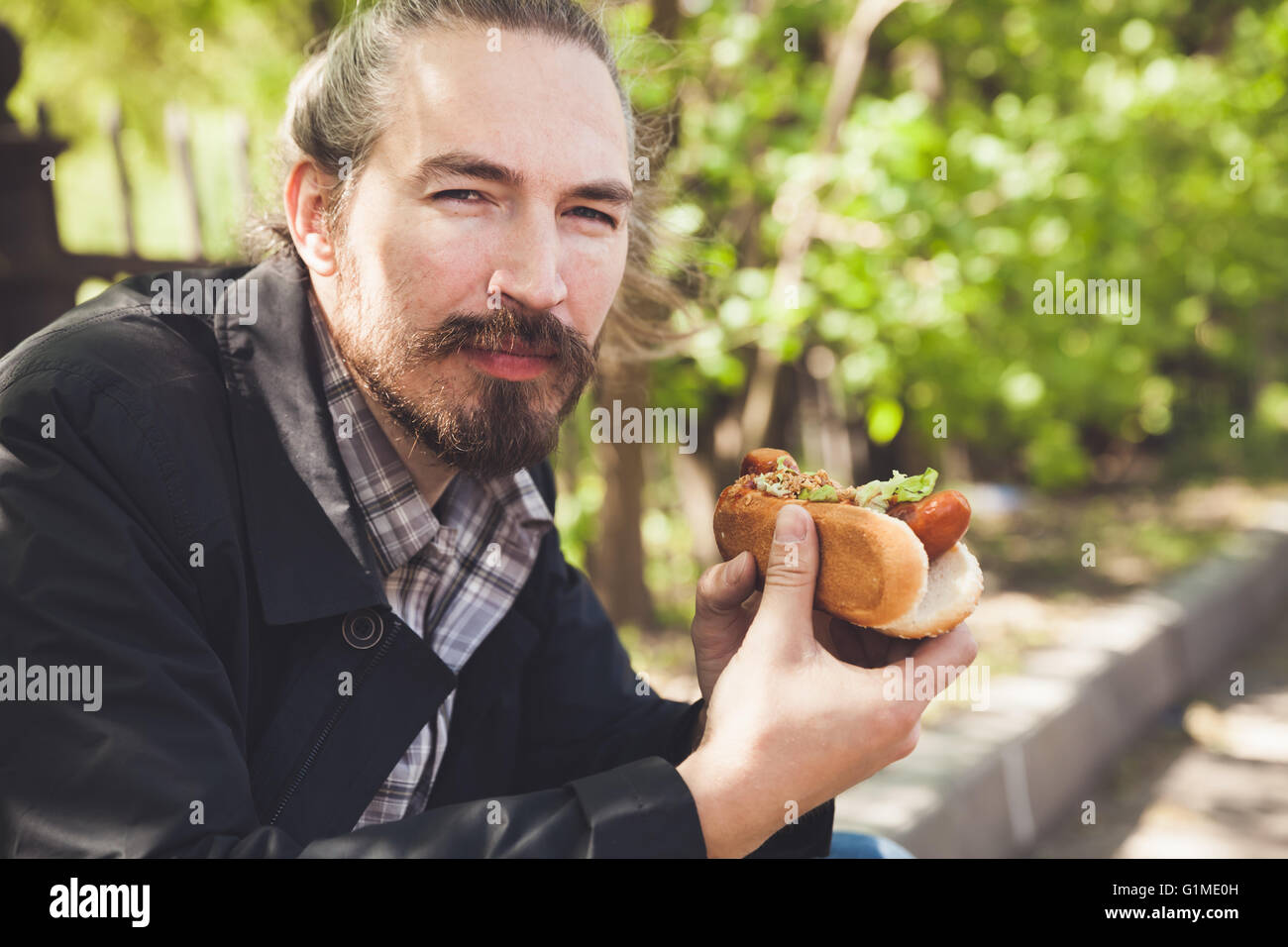 Bärtige asiatischen Mann mit Hot-Dog hat eine Mittagessen im Sommerpark, Outdoor-Portrait mit selektiven Fokus Stockfoto