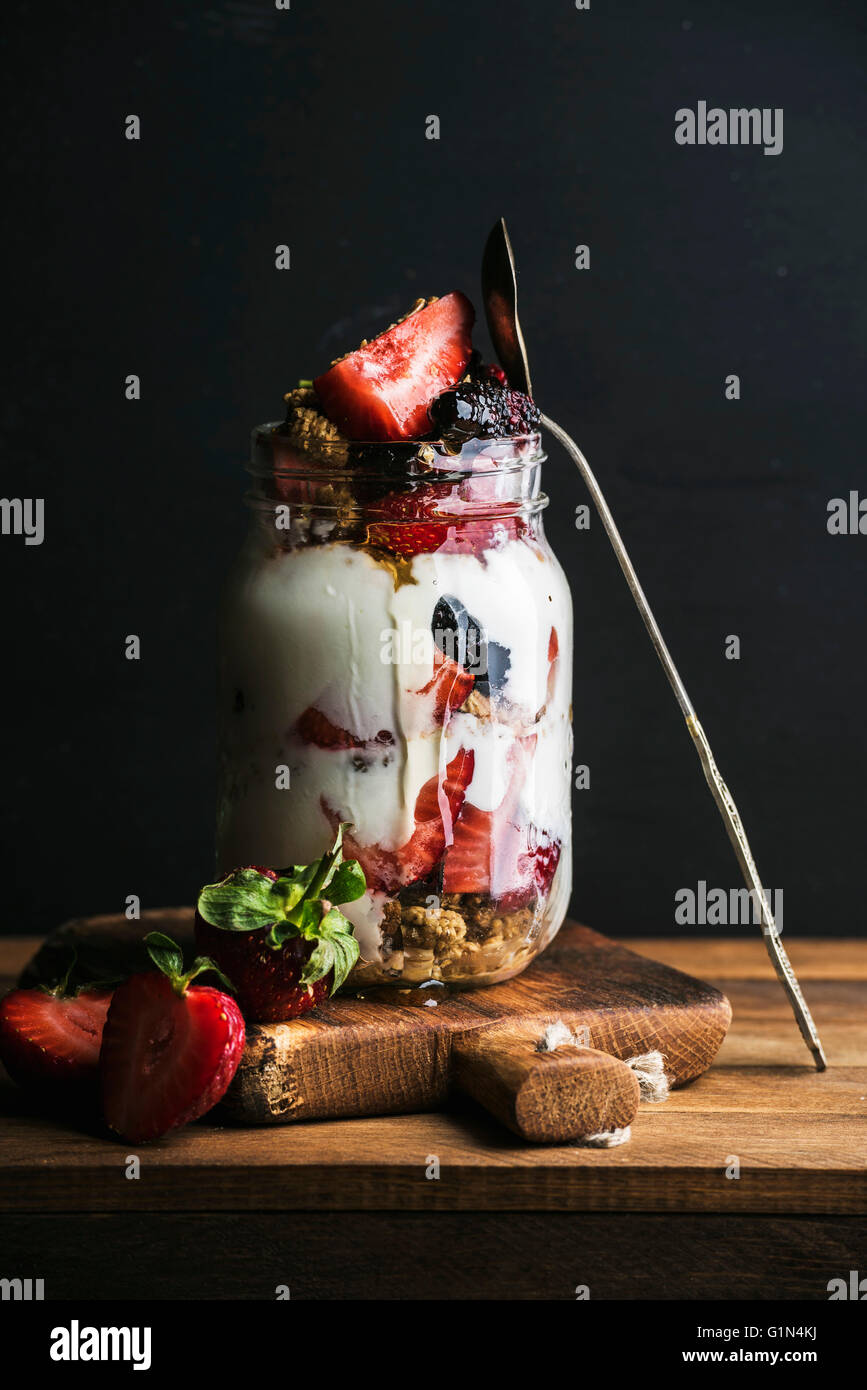 Joghurt-Hafer-Müsli mit Erdbeeren, Maulbeeren, Honig und Minze Blätter in Longdrinkglas Glas auf schwarzem Hintergrund Stockfoto
