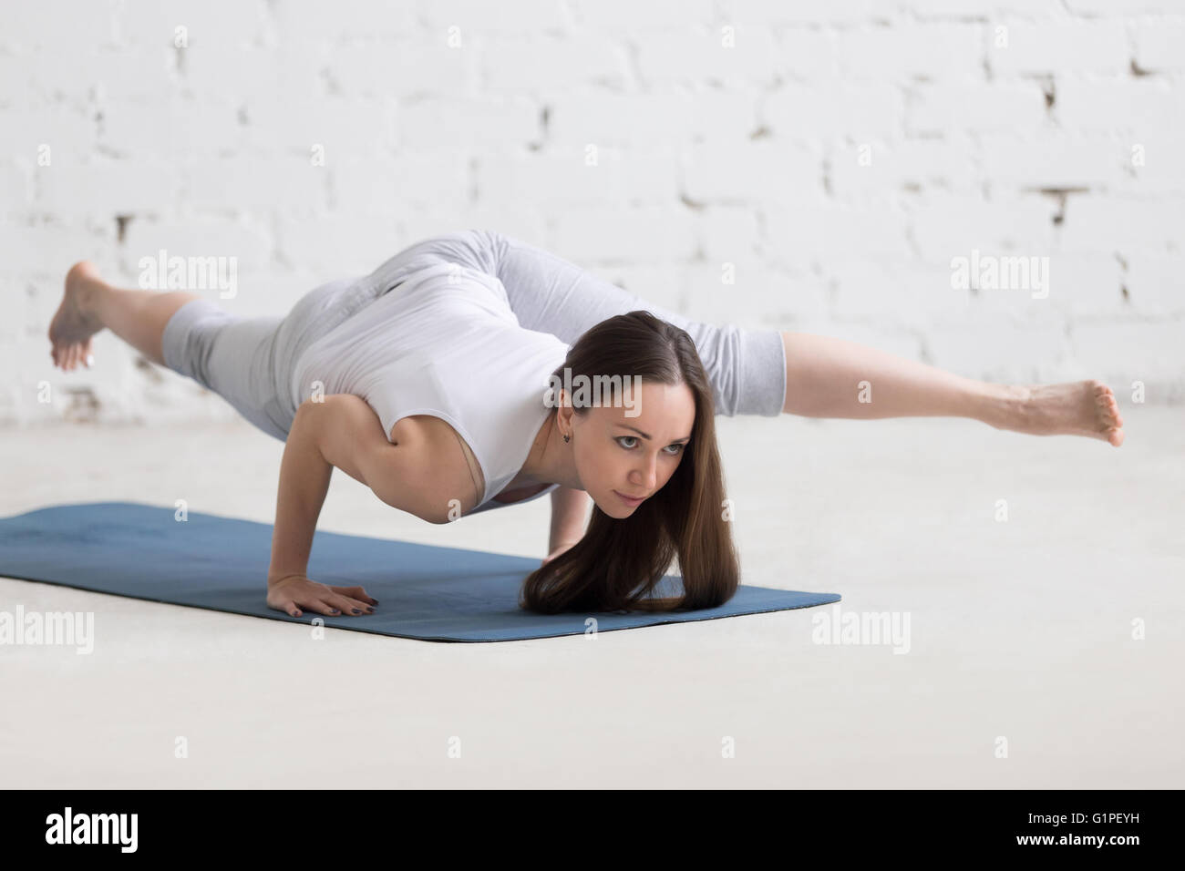 Attraktive junge Frau trainieren. Schönes Modell Übungen auf blauen Matte im Raum mit weißen Wänden. Eka Pada Koundinyasana Stockfoto