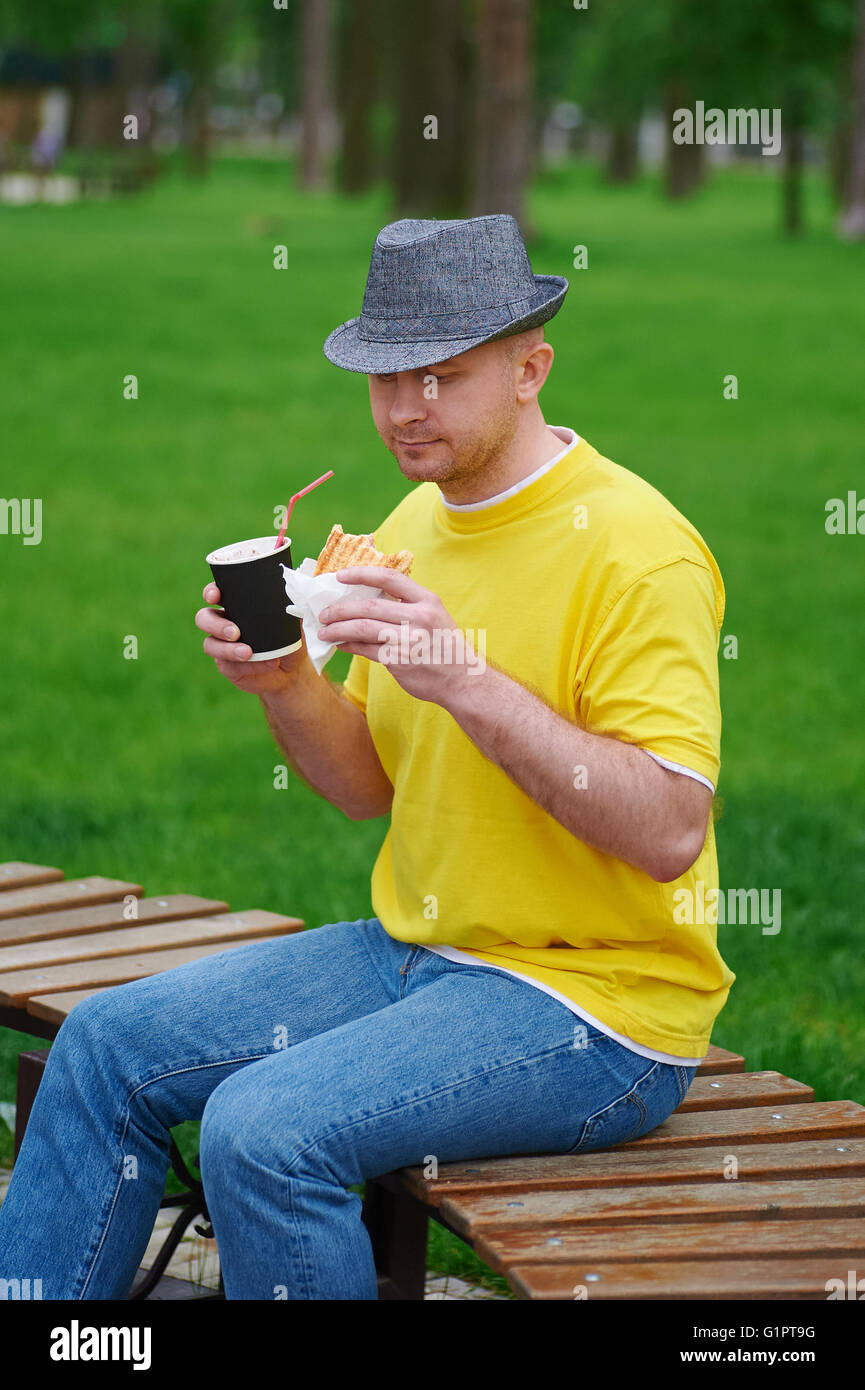 Hübscher junger Mann sitzt im Park auf Holzbank Lächeln und genießen einen Kaffee Stockfoto