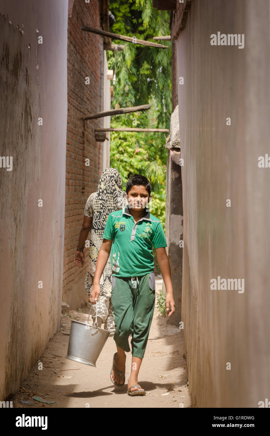 Kleiner Junge geht auf eine Gasse trägt einen Eimer mit Wasser in einem Dorf in Gujarat, Indien Stockfoto