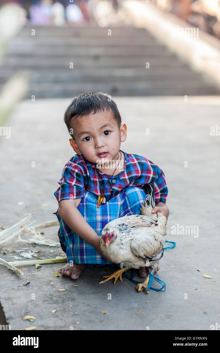 Ein burmesisches Kind aus Mandalay, Burma, Myanmar, schaut direkt in die Kamera, hockt und hält ein weißes Huhn in den Händen. Stockfoto