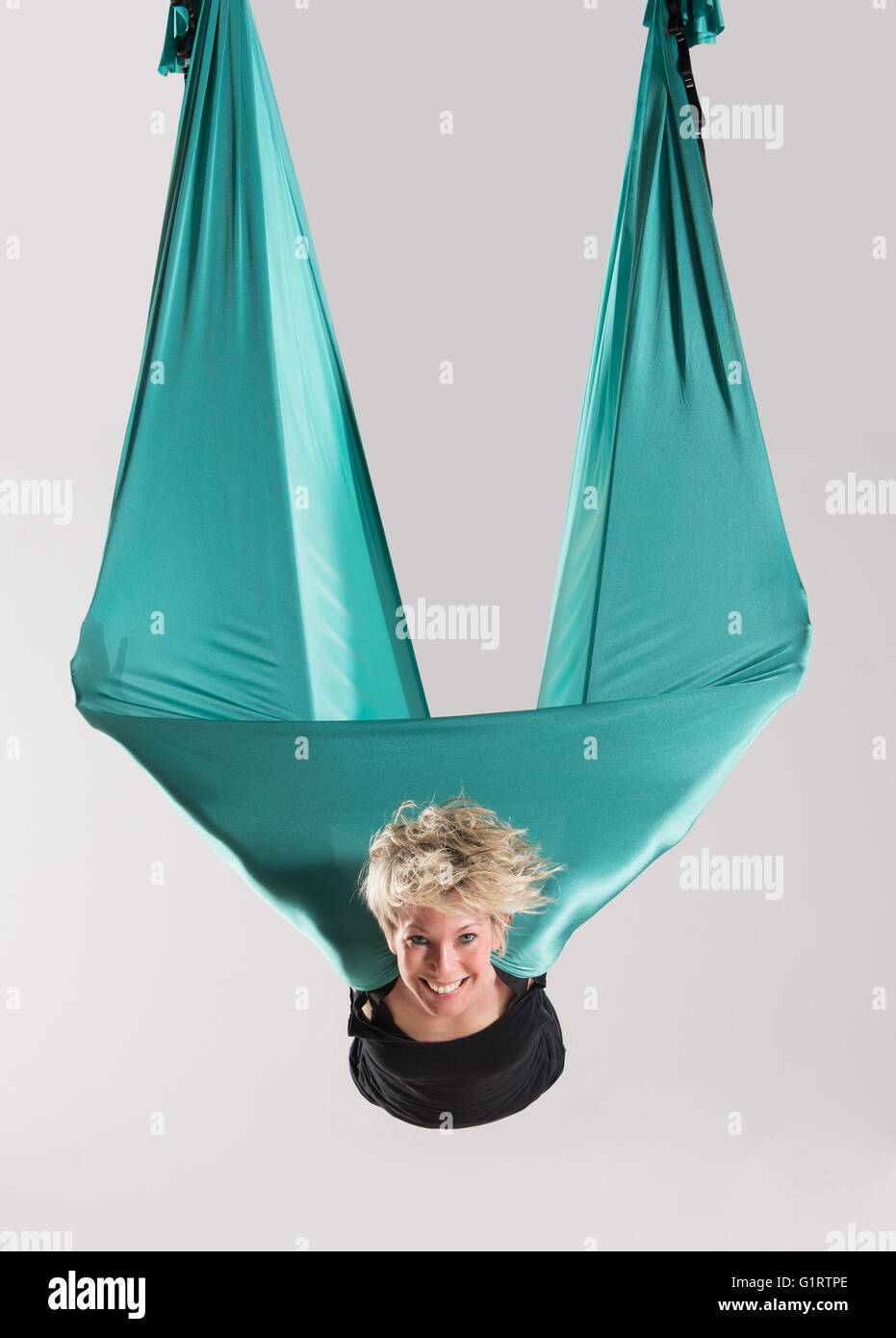 Lächelnd Mitte Luft junge blonde Frau aerial Yoga zu tun, mit Arme in großen grünen flexible Tarp abgehängte Decke gewickelt Stockfoto