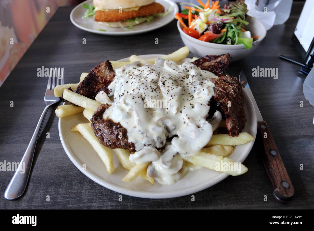 Ein Teller mit Steak mit Pilzsauce und Pommes Frites mit einer Schüssel griechischen Salat und Huhn Schnitzel Burger im Hintergrund Stockfoto