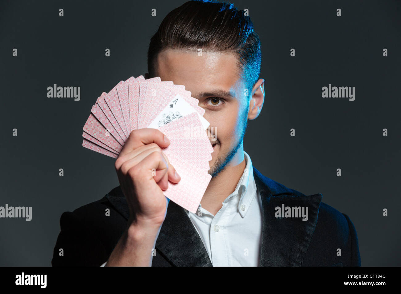 Lächelnde junge Zauberer bedeckte sein Gesicht mit Spielkarten auf grauem Hintergrund Stockfoto