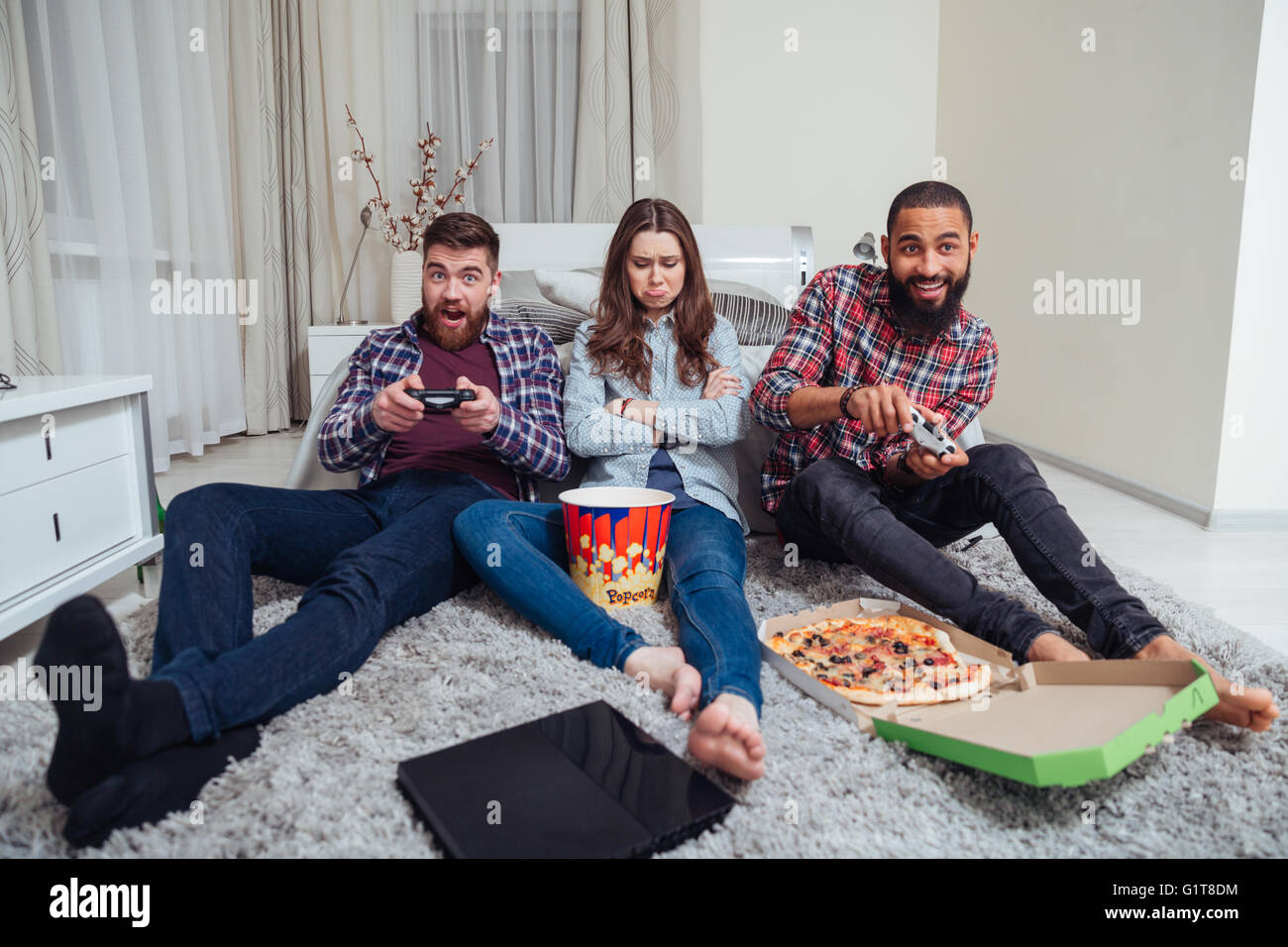 Zwei glückliche junge Männer spielen Computerspiele und traurig beleidigt junge Frau sitzt auf dem Zimmer Stockfoto