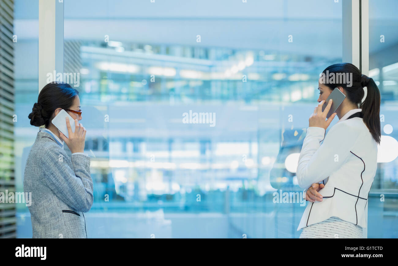 Geschäftsfrauen auf Handys am Bürofenster im Gespräch Stockfoto