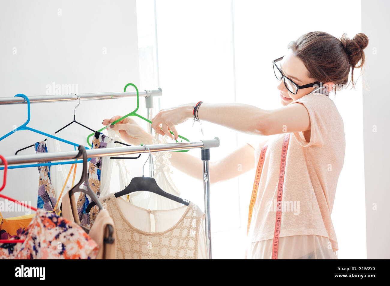 Lächelnde hübsche junge Frau-Mode-Designer in Gläsern prüfen neue Sammlung von Kleidung und reden über Handy Stockfoto