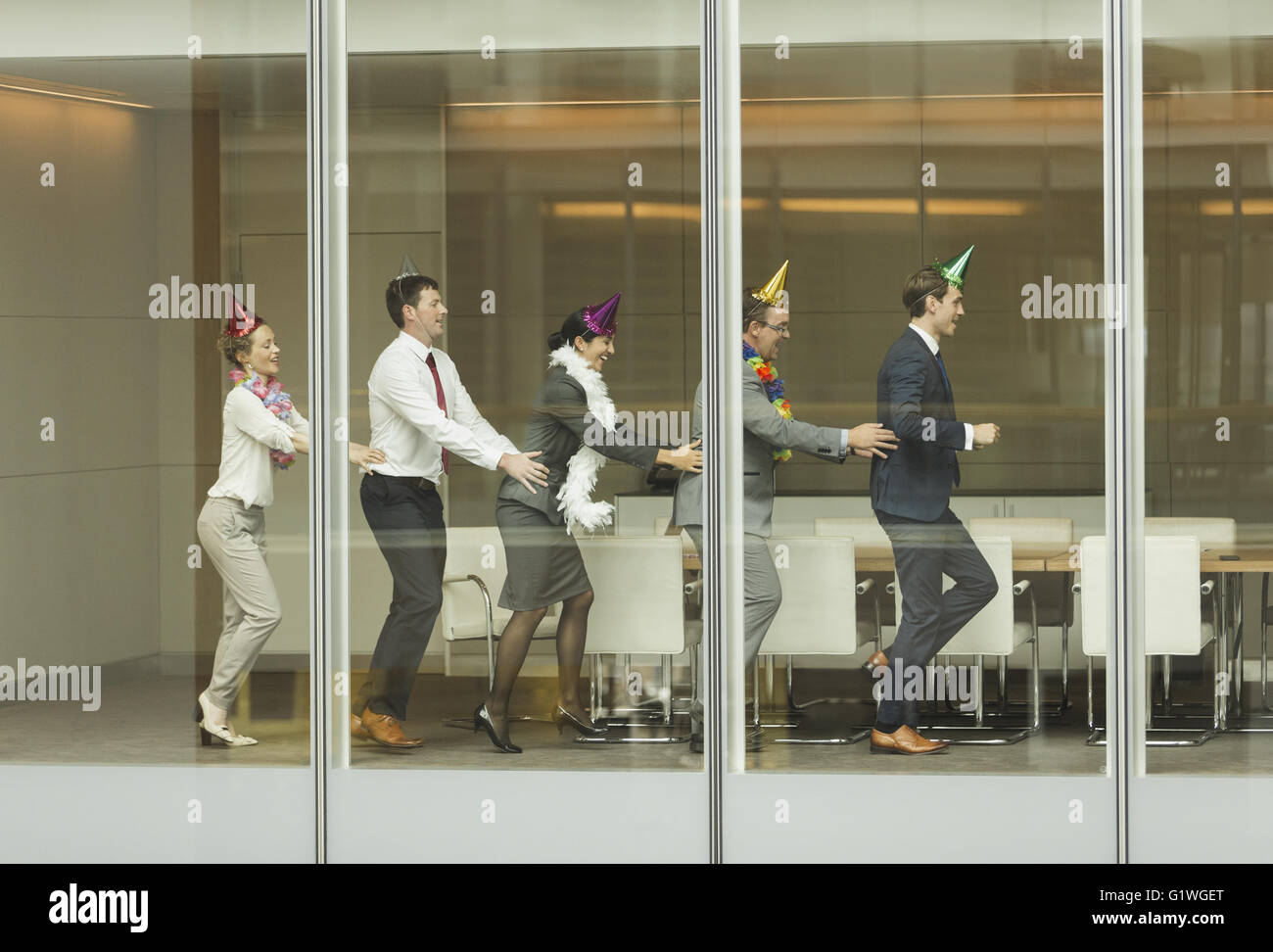 Business-Leute tragen Partyhüte tanzen in Conga Linie an Konferenz Zimmer Fenster Stockfoto