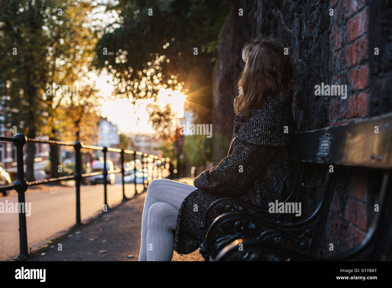 Eine junge Frau sitzt auf einer Bank bei Sonnenuntergang an einem Herbsttag in der Stadt Stockfoto