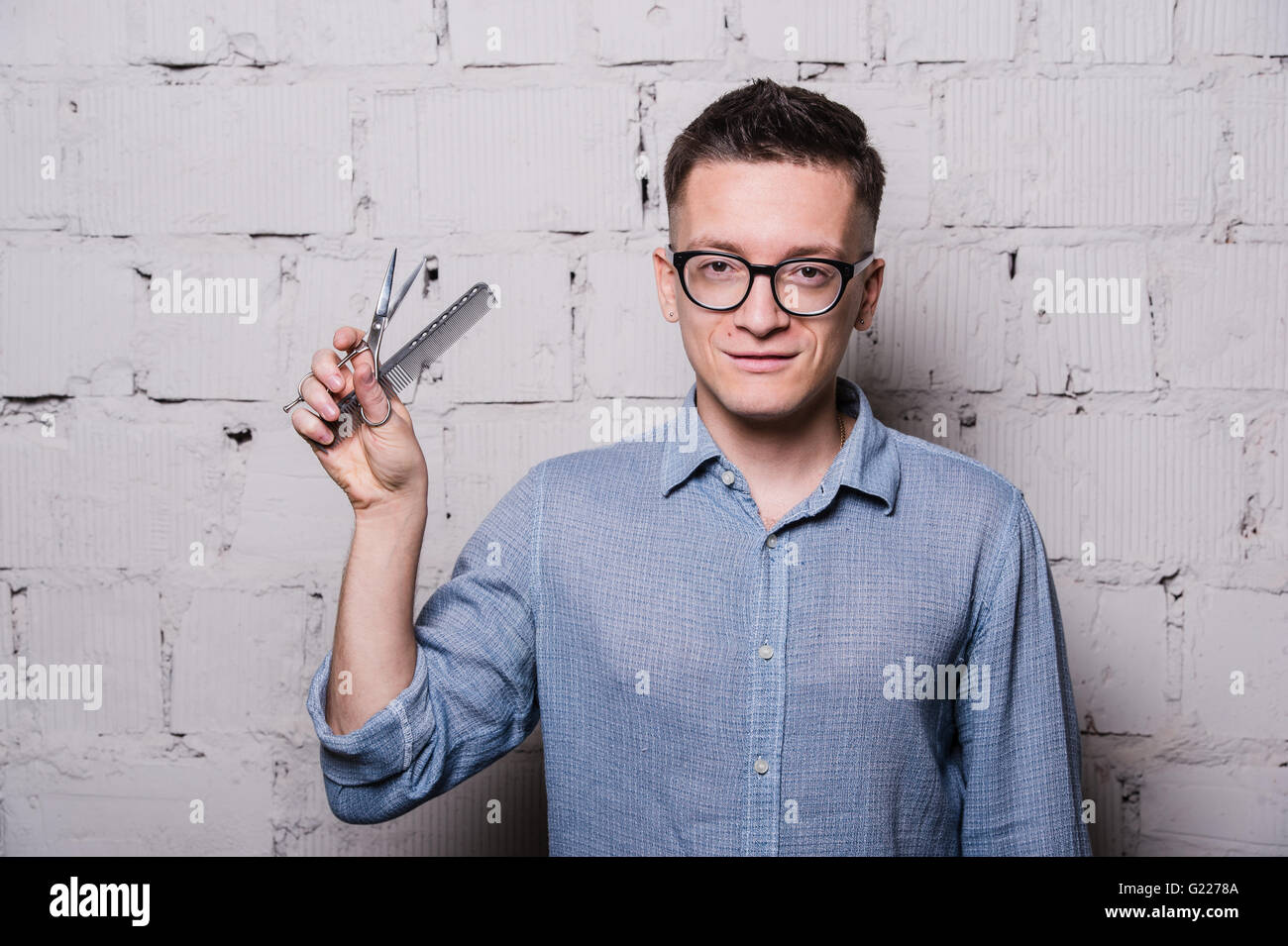 Hübscher Junge männliche Friseur in Gläsern, posiert mit Schere und Kamm, auf grauen Ziegel Wand Hintergrund, horizontales Bild Stockfoto