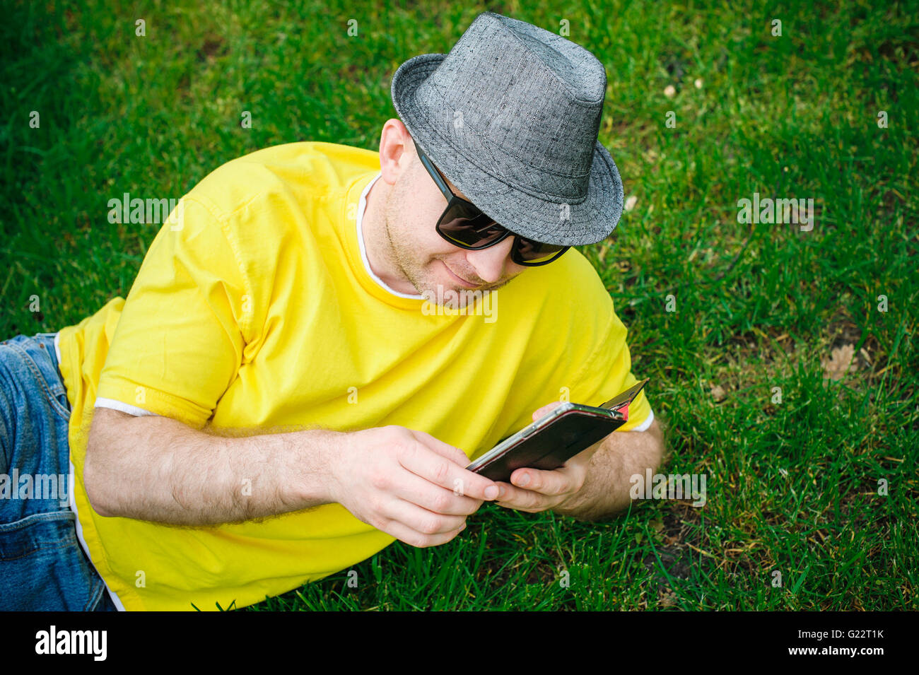 Porträt der jungen lächelnd Mann in Hut und Brille sitzt auf dem grünen Rasen mit Telefon Stockfoto